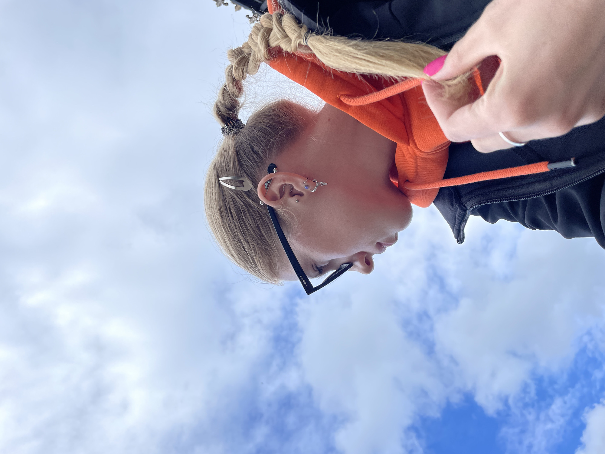 The image, presented sideways, captures a close-up, low-angle view of a person's head and upper body against a bright sky backdrop. The person, with blonde hair styled in two thick braids, wears an orange hooded sweatshirt under a dark jacket. The visible left ear is adorned with numerous and varied piercings, including those on the tragus, daith, conch, and multiple helix piercings, one of which is a decorative barbell along the upper cartilage. A silver hair clip is also seen on the side of the head, near the ear. Black-framed glasses rest on the bridge of their nose, with the text 'VOGUE' clearly visible on the temple arm. A right hand, featuring pink nail polish and a silver ring, is shown gently touching a strand of hair from one of the braids. The background consists of a bright blue sky with scattered white, fluffy clouds, indicating a daytime outdoor setting. This scene is located in Izhevsk, Russia.
