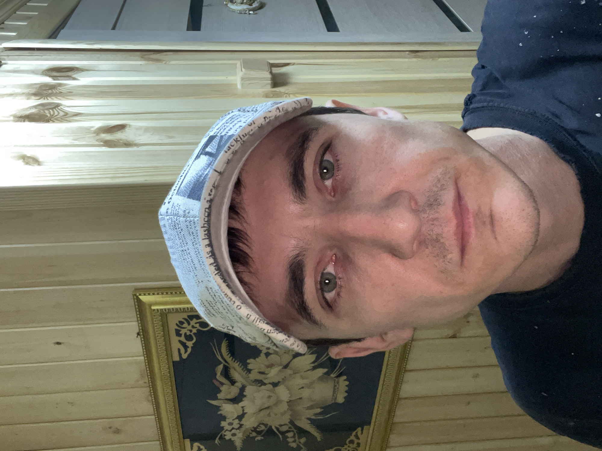 The image is a close-up, selfie-style photograph of a man wearing a light blue baseball cap and a dark t-shirt. He has green eyes and a slight mustache. The background consists of wooden paneled walls and a framed picture on the left. The photograph does not provide any specific details that would allow for its location to be identified as Laishevo, Russia.
