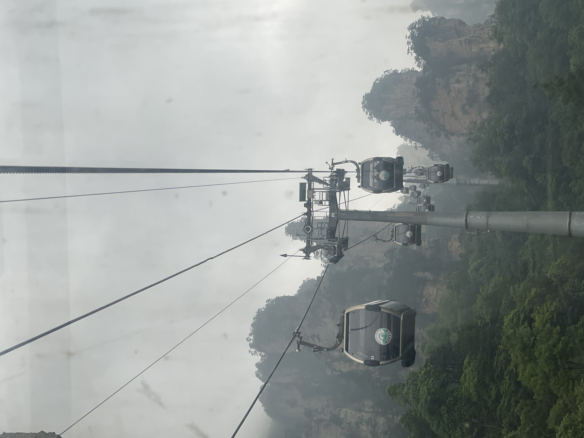 The image depicts a cable car system in Wulingyuan, China, shrouded in mist. The cable cars are modern and enclosed. The landscape consists of tall, rocky pillars covered in vegetation and obscured by fog. The scene is vertically oriented, as the picture was taken and saved with the phone sideways. The cables are taut and connect to supporting towers.