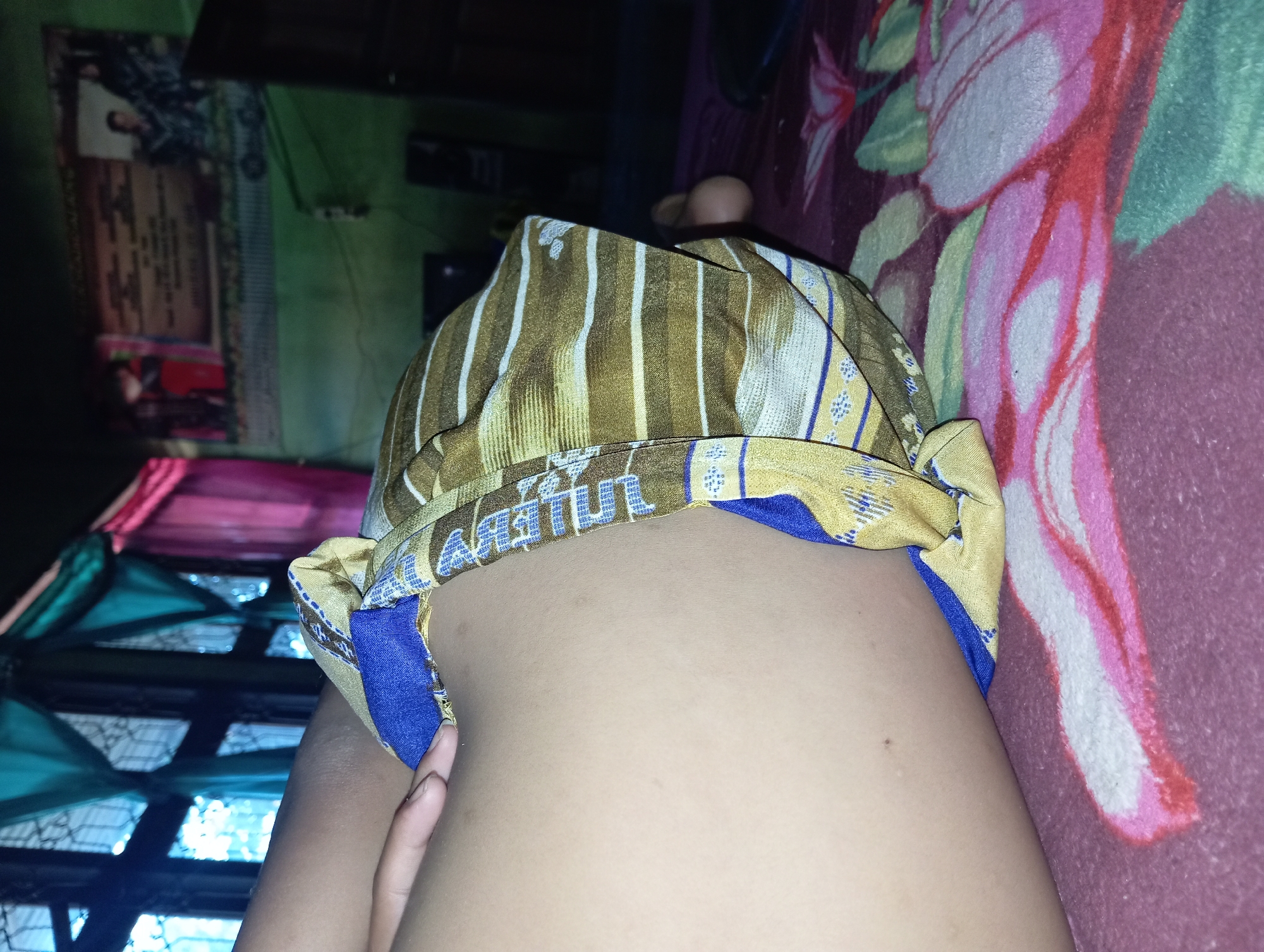 The image depicts a close-up, low-angle shot of a person's midsection, covered by a patterned cloth, likely a sarong. The sarong features stripes of gold, white, and blue with a repeating blue floral motif. The person's skin, visible on the left side of the frame, appears light-toned and smooth, with a small, dark mole or blemish visible. The sarong is draped over the person's body and rests on a blanket with a large floral pattern in shades of pink, white, and green.
The background is dimly lit and appears to be an indoor setting, potentially a bedroom. To the left, blue and turquoise curtains are visible, framing a window. Above the person's midsection, a poster or banner is partially visible, featuring text and images, though they are out of focus. The overall impression is one of a domestic environment, possibly in Rejosari, Indonesia, given the context. The lighting is dim, suggesting it may be evening or nighttime, or that the room is not well-lit. The focus is tight on the person's body and the sarong, with the background elements providing context but not being the primary subject.