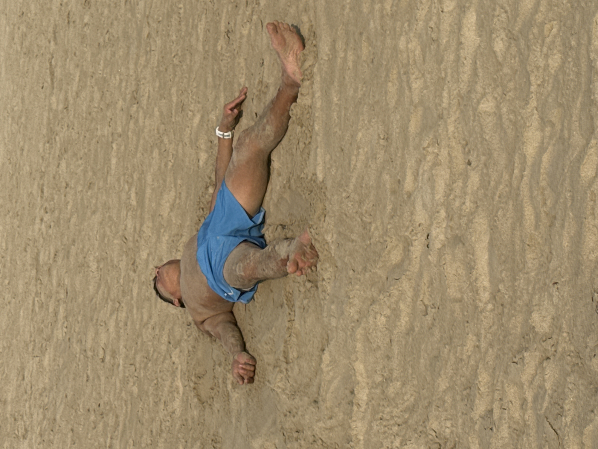 An aerial view shows a man in blue swim trunks lying face down on a beach. His left arm is extended upward, with his hand open, and his right arm is bent at the elbow, with his fist clenched. His legs are also extended upward, with his toes pointing up. A white watch is visible on his left wrist. The sand is light brown and appears damp, with various footprints and patterns visible. The location is likely a beach in Đà Nẵng, Vietnam, based on the sandy environment.
