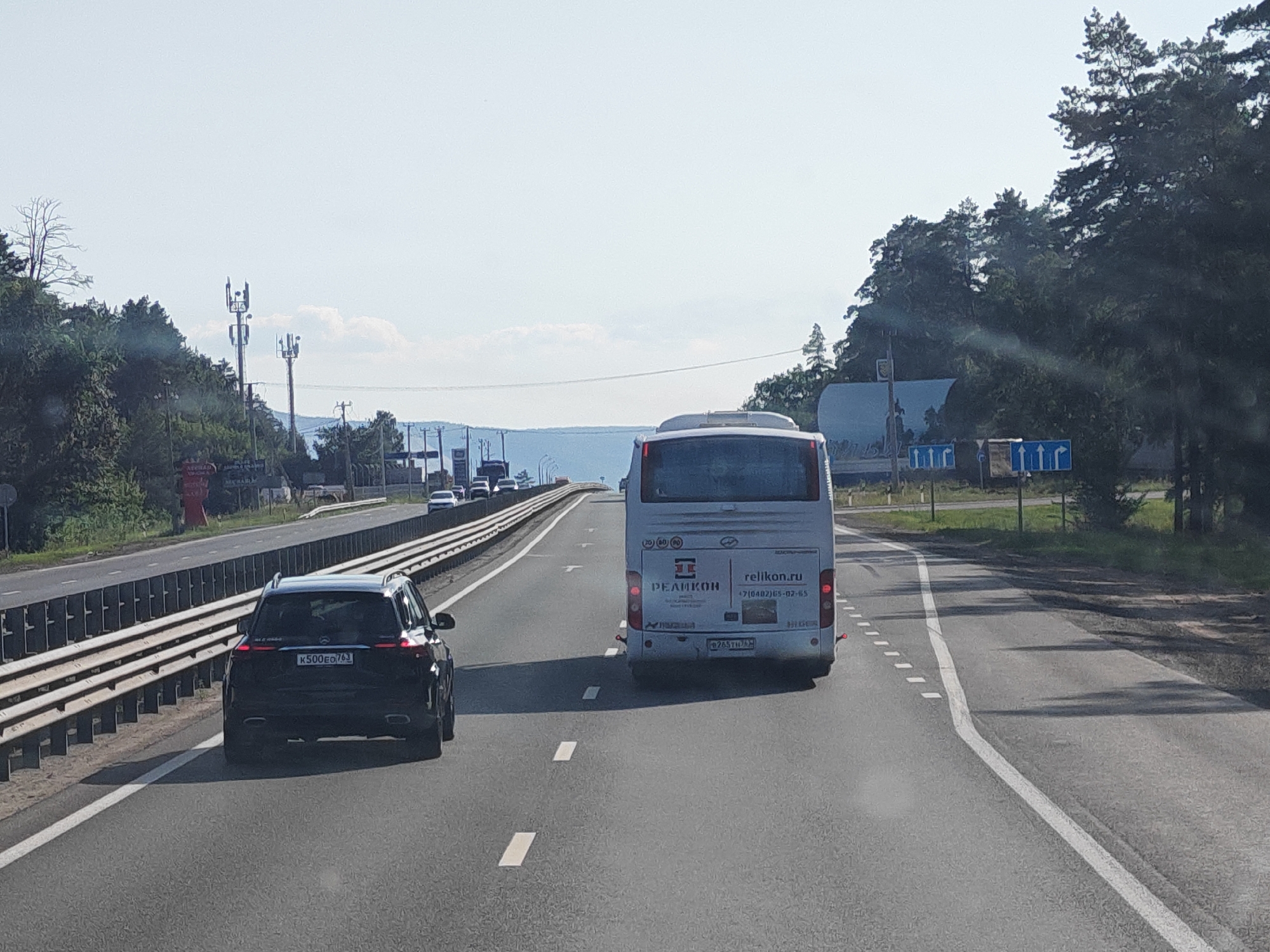 The image depicts a view from a vehicle traveling on a highway. In the foreground, a black SUV is in the left lane, and a white bus is in the right lane. Both vehicles are moving forward. The highway has multiple lanes, divided by white lines. A metal guardrail runs along the left side of the highway. Trees and vegetation line the sides of the road, and in the distance, hills and a body of water are visible. The sky is clear and bright. There are traffic signs visible in the background, including speed limit indicators and directional arrows. The scene appears to be set on a sunny day. The bus has branding for "RELIKON.RU" and a phone number. The license plate of the SUV is partially visible. The image does not contain any explicit elements to confirm the exact location within Rassvet, Russia, but it shows a typical highway scene in a populated area with natural surroundings.