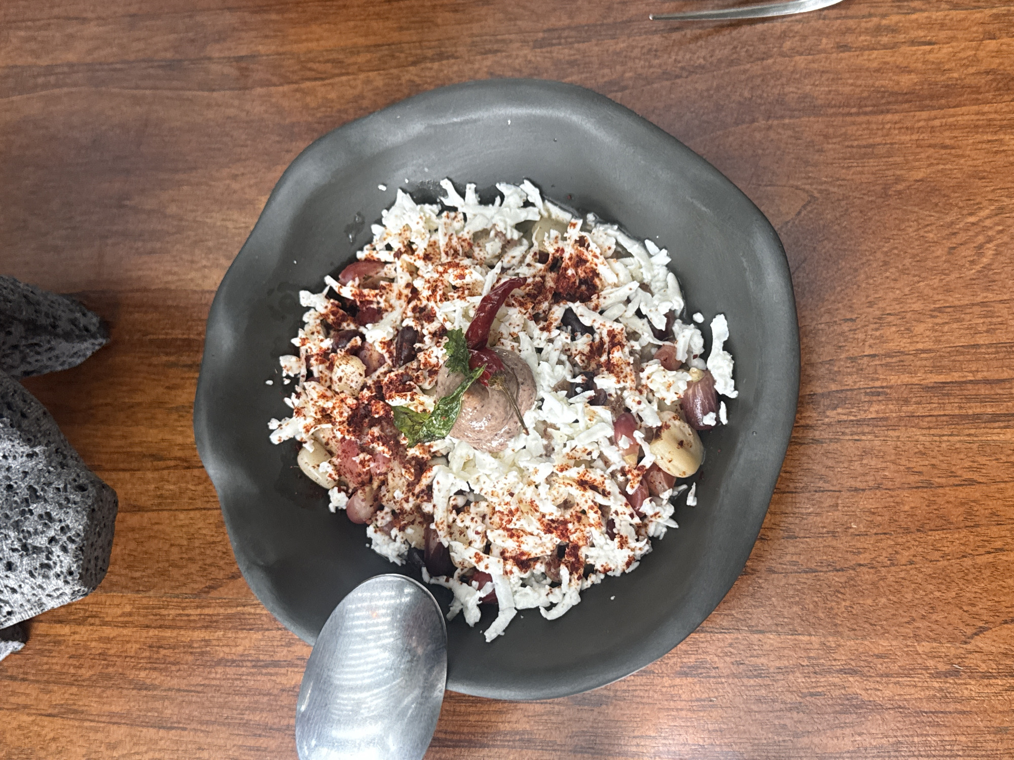 The image shows a plate of food on a wooden table. The plate is a dark grey, irregular shape, and it contains a dish with shredded white cheese, beans, some sort of pale round bean or vegetable, and a reddish-brown spice sprinkled on top. There are also some small green herbs or sprouts on the plate. A spoon is positioned next to the plate, and a textured grey object is partially visible on the left side of the frame. The photo was likely taken in Mexico City, Mexico, given that the food served contains similar ingredients to the regional cuisine.