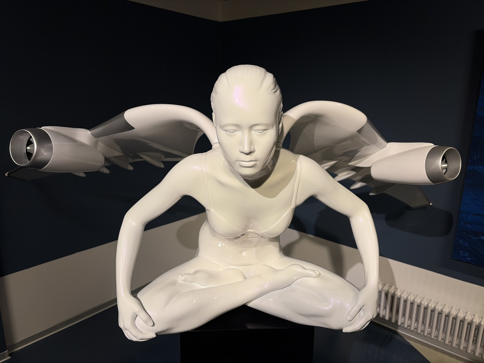 A white, glossy sculpture of a seated female figure with wings is displayed against a dark blue background. The figure has its legs crossed in a meditative pose, and its arms are bent at the elbows with hands resting on its knees. The wings appear to be designed to resemble airplane engines, with large cylindrical nacelles attached to sleek, aerodynamic surfaces. The sculpture is illuminated from the front, casting shadows on the background. The environment includes a radiator visible at the bottom right of the frame. The sculpture is situated in the Saint Petersburg Kunstkamera Museum.