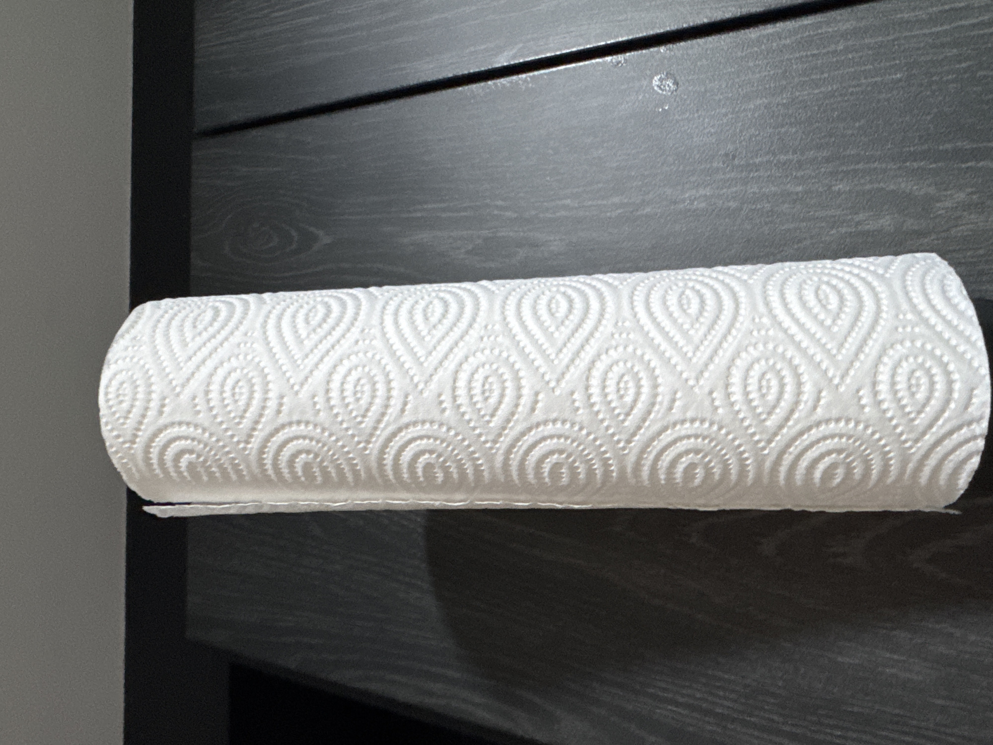 A single, full roll of white paper towels stands vertically against a dark gray textured background. The paper towels feature an embossed pattern of interconnected swirling ovals and smaller dotted teardrop shapes. The dark gray background surface, possibly wood or laminate, exhibits faint vertical grain lines. A distinct, soft shadow from the left side of the paper towel roll is cast onto this surface, indicating a light source from the right. A thin, darker horizontal edge separates the main dark background from a lighter gray surface above it. To the right of the paper towel roll, the dark background surface is interrupted by a vertical, shiny black line or seam, separating two panels. Small, reflective specks are visible on the rightmost panel near this seam. There are no people present, and no identifiable markings or elements indicating a specific location in Bangkok, Thailand, or any other city.