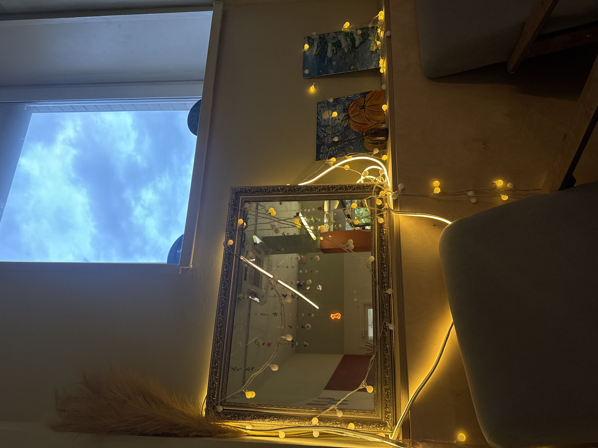 In an indoor setting, likely a room or cafe, a framed mirror with ornate gold detailing sits on a shelf. The mirror is adorned with a string of warm yellow fairy lights, some of which are circular and others are bulb-shaped. Reflected in the mirror is a room with hanging decorations and a neon orange '1' sign. To the right of the mirror, also on the shelf, are two small paintings: one depicts three orange pumpkins under a starry night sky, and the other shows a snow-covered Christmas tree. The shelf is illuminated by the fairy lights and a bright light source, possibly a ceiling light or natural light from a window above. The window, visible in the upper portion of the image, looks out onto a cloudy blue sky. A tall, feathery dried plant is positioned to the left of the mirror. In the foreground, the backs of two light-colored armchairs are visible, suggesting a seating area. The overall ambiance is cozy and decorative, with a warm glow from the lights.