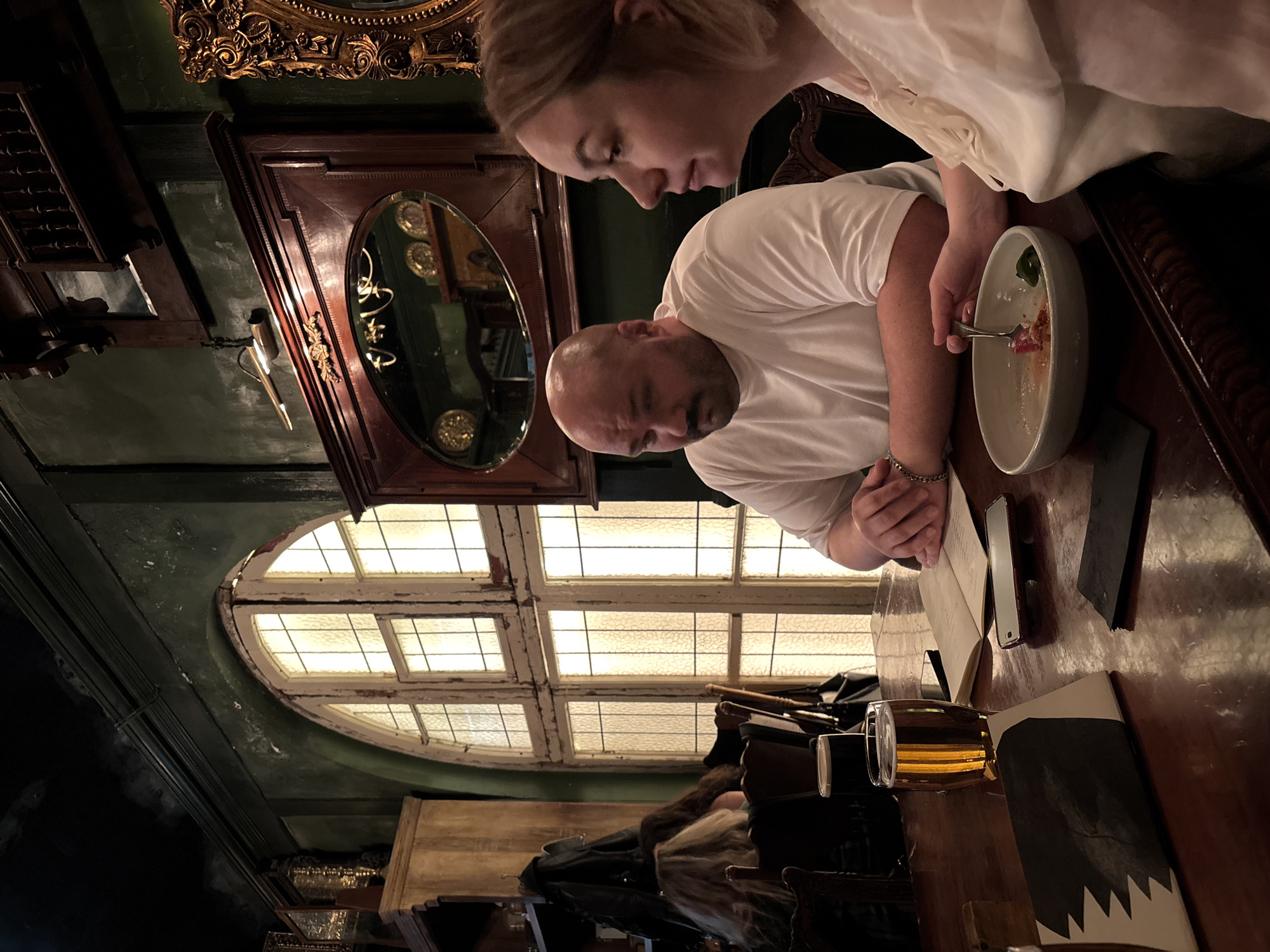 A medium shot depicts a man and a woman seated at a dark wood table in what appears to be a restaurant. The man, who is bald, is wearing a white t-shirt and is looking down into a partially eaten bowl of food, holding a fork in his right hand. The woman, also wearing a white shirt, is leaning in towards the bowl. On the table, there's a glass of amber-colored liquid, possibly beer, along with a menu and what appears to be a smartphone and possibly a knife. The setting features an arched window with a multi-paned design and ornate wooden detailing in the background. A dark-colored wall is visible, with a framed mirror and other decorative elements. The presence of such items and the architectural features suggest a potentially historic or upscale establishment. There is not enough information in the image to pinpoint the location in the city of Moscow, Russia.
