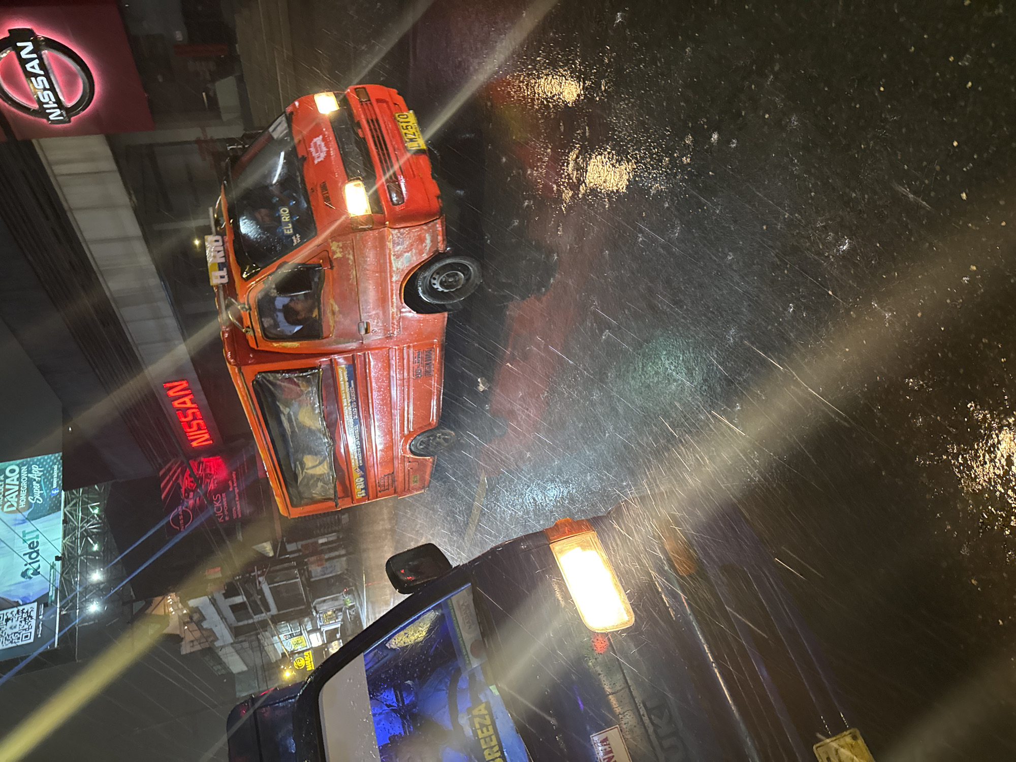 The image depicts a rainy night scene in Davao City, Philippines. A red multicab, a common form of public transportation in the Philippines, is driving on a wet street, reflecting the headlights and streetlights. The multicab has "EL RIO" written on its roof and the license plate reads "LWZ 510". In the foreground, the front of a dark-colored vehicle is visible, also illuminated by its headlights. In the background, illuminated signage for Nissan is prominent, along with other businesses and a large digital billboard displaying "RidelT DAVAO HOMEGROWN Super App". The wet asphalt of the road and the streaks of rain indicate heavy precipitation. The overall impression is of a busy urban environment during a downpour.