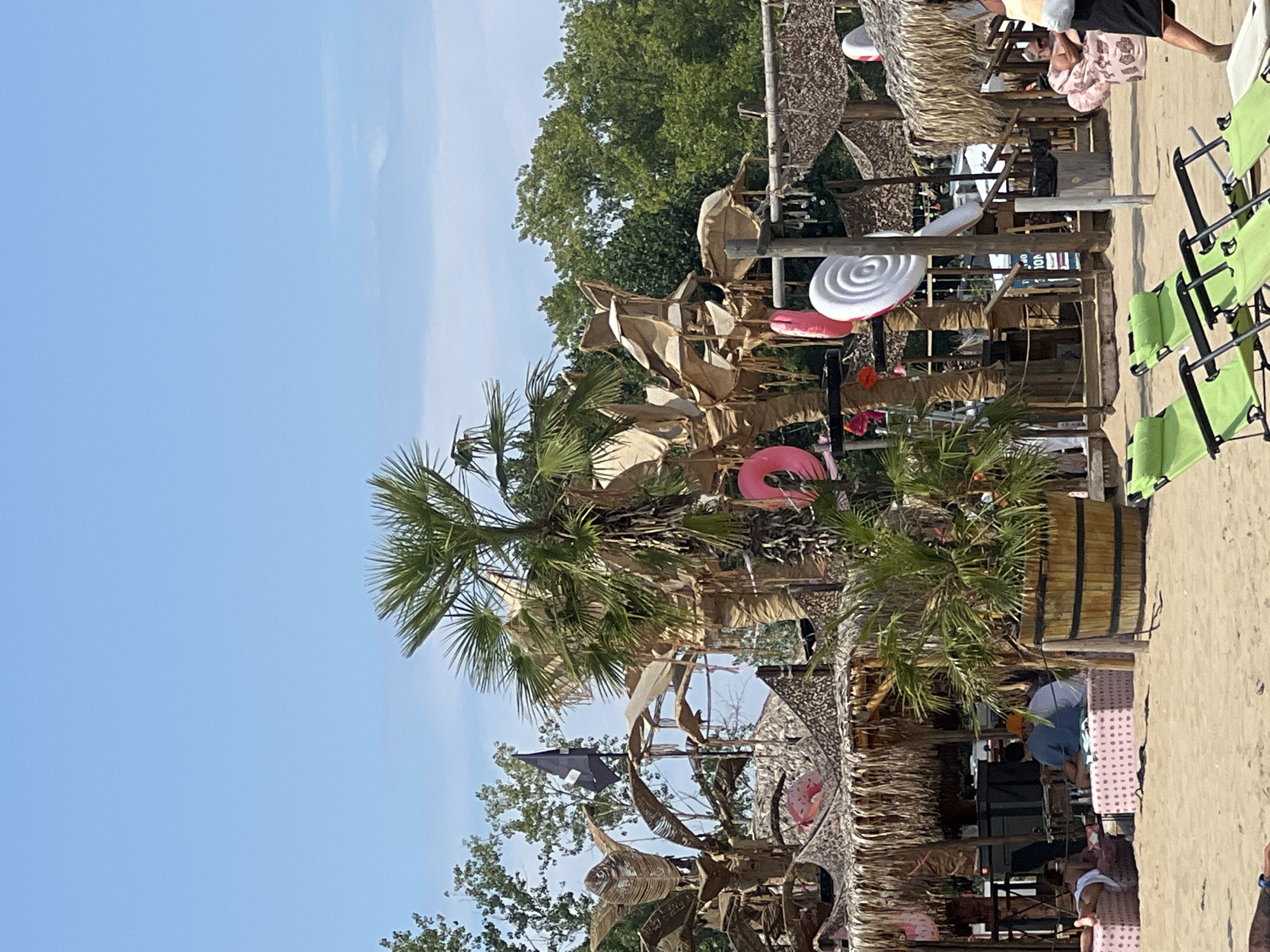 The image shows a beach scene with several palm trees decorated with inflatable rings and lollipops. There are thatched-roof structures that appear to be bars or seating areas. People are visible in the background, some sitting on lounge chairs or at tables. The overall ambiance suggests a relaxed, tropical-themed environment. The specific location in Sozopol, Bulgaria, is not identifiable from the image alone.