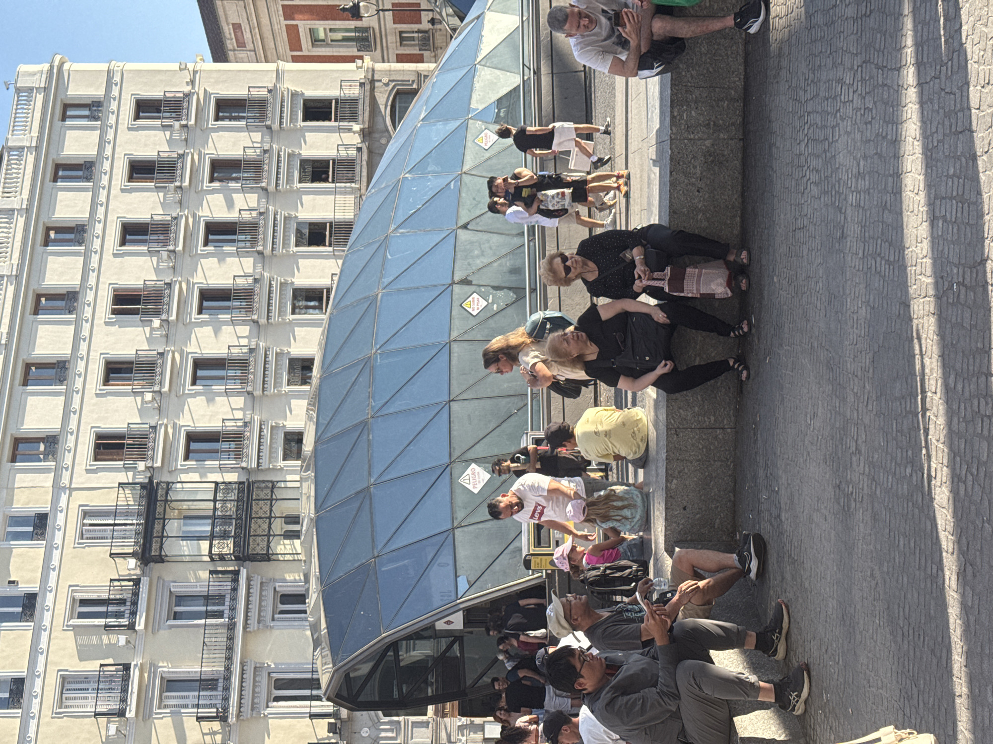 The image depicts a street scene in Madrid, Spain, likely in the Puerta del Sol area, given the distinctive glass pyramid structure, which is an entrance to the Sol metro station. The background features a large, light-colored building with multiple floors, adorned with numerous windows and balconies, a common architectural style in Madrid. In the foreground, several people are seated on a stone ledge.
On the left, a man wearing glasses and a grey jacket is looking down at his phone, with a bottle of water next to him. Beside him, another man in a grey t-shirt and a cap is also looking at his phone. A young girl in a pink hat and a patterned dress is sitting with her back to the camera. Further to the right, a woman in a black top and sunglasses is seated with a patterned bag on her lap. Next to her, another woman in a black polka-dot top and dark pants holds a large pink and white patterned tote bag. To the far right, a man in a white t-shirt and black shorts is looking at his phone.
Behind the seated individuals, more people are visible, some walking and others standing. A man in a white t-shirt with "LEVIS" written on it is interacting with a young girl. The glass pyramid structure is prominent, with its geometric panels reflecting the sky and surroundings. Signs, including a triangular warning sign with an exclamation mark and the word "PELIGRO" (danger), are visible on the pyramid. The ground is paved with cobblestones, and shadows are cast from the building and the pyramid, indicating a sunny day. The overall atmosphere appears casual and public, with people engaged in various activities typical of a city square.
