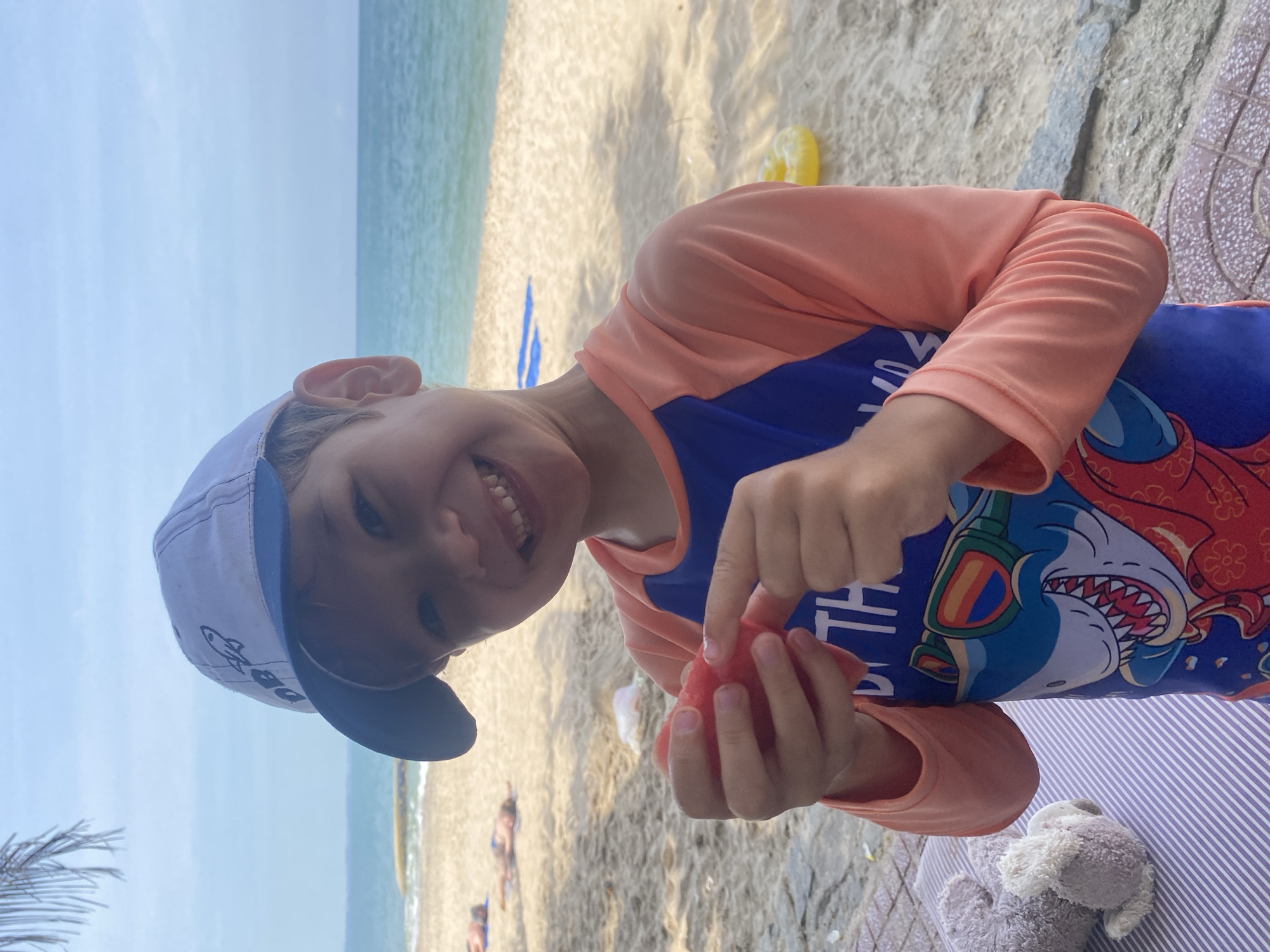 A young boy wearing a blue and orange rash guard and a white baseball cap with a rocket on it is holding a piece of red fruit, possibly watermelon, and smiling at the camera. He is on a sandy beach with the ocean in the background. Several people are lying on towels on the beach in the distance. A yellow inflatable ring is visible on the sand to the right of the boy, and a stuffed animal is on a striped mat to the left. The sky is a pale blue with scattered clouds.