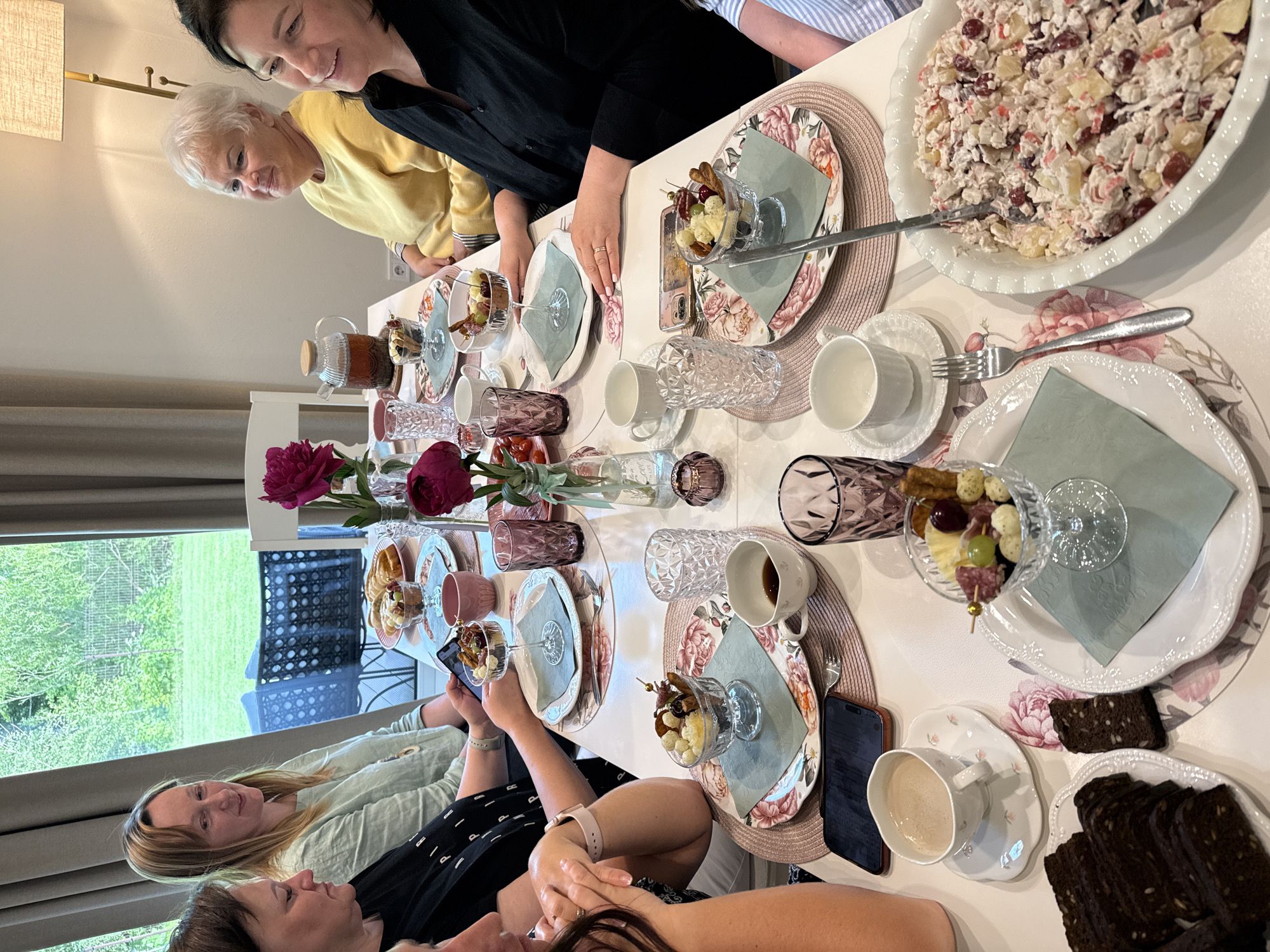 A group of seven women are seated around a table indoors for what appears to be a brunch or tea gathering. The table is set with floral-patterned placemats and plates, various dishes of food, and drinks. The women are casually dressed and engaged in conversation. One woman in the foreground is looking at her phone. The room has a large window with a view of greenery and what appears to be outdoor furniture. The setting suggests a home or a small cafe. The location is Rāmava, Latvia, as indicated by a watermark on the original image.