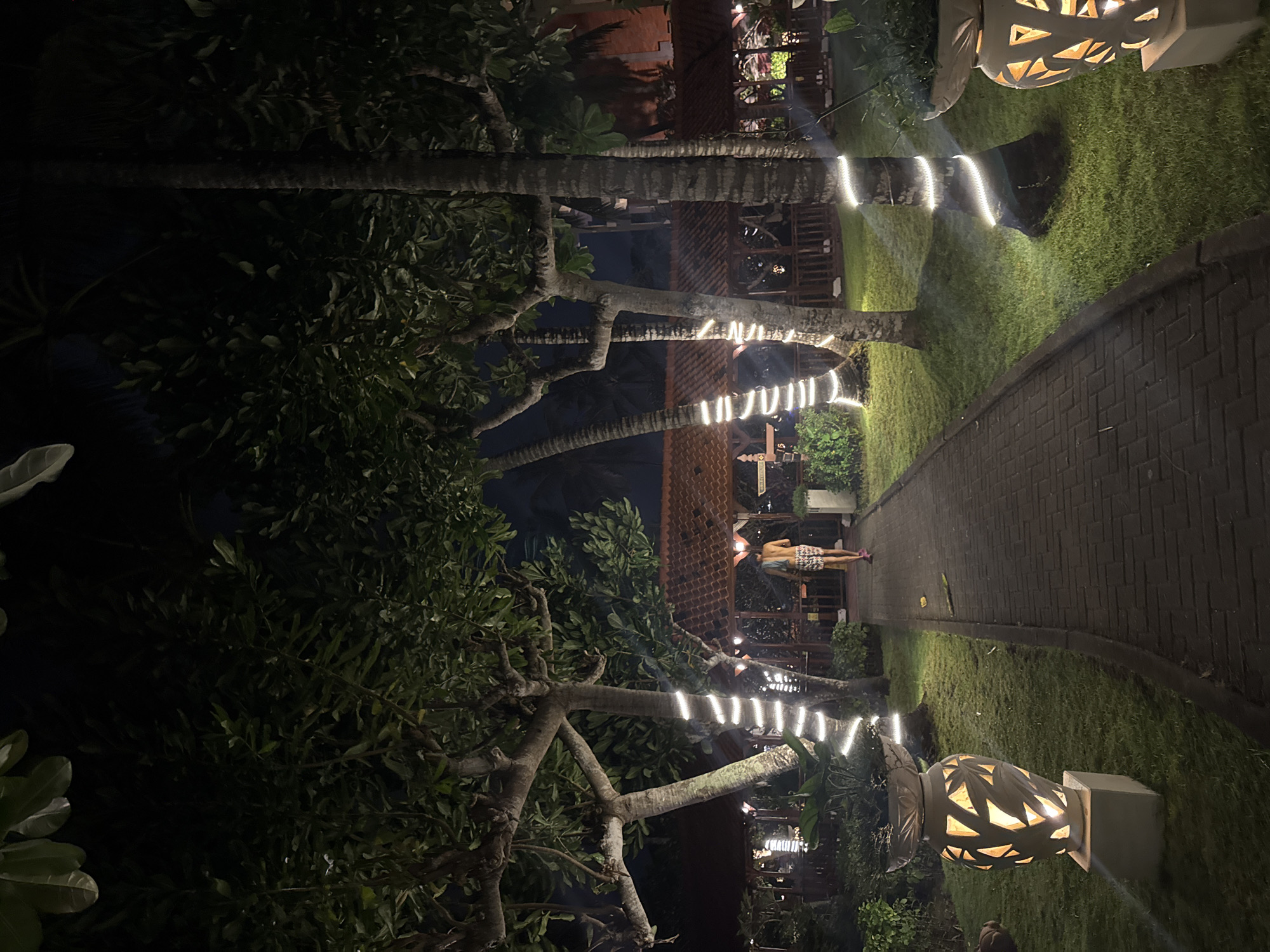 The image depicts a pathway at night in Kuta Selatan, Indonesia, possibly within a resort or restaurant complex, illuminated by decorative white string lights wrapped around palm and other trees. The pathway is paved with dark bricks and bordered by well-maintained green grass. On either side of the pathway, large decorative urns with intricate cut-out patterns are lit from within, casting warm light onto the grass. In the background, structures with reddish-brown tiled roofs and wooden railings suggest an outdoor dining or seating area, also decorated with lights. A lone individual, viewed from behind, walks away from the camera down the pathway, wearing patterned shorts and a light-colored top, with a towel or cloth slung over their shoulder. The overall atmosphere is one of tranquil evening ambiance in a tropical setting.