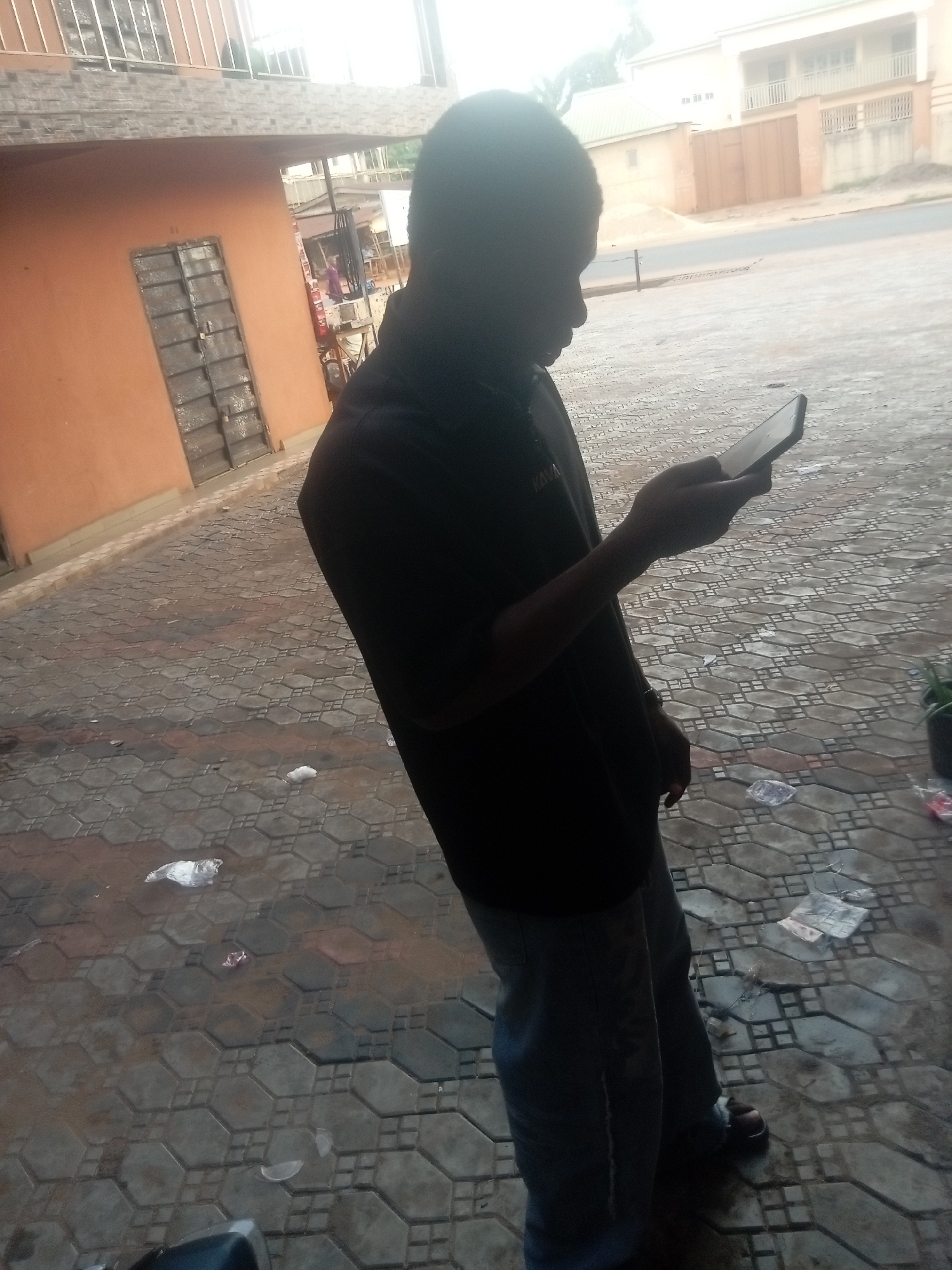 A person stands on a paved area, looking at a mobile phone. The person is wearing a dark shirt and light pants with sandals. The background shows a building with an orange facade and a metal door to the left, and a building with a lighter exterior across a street in the distance. The paving stones are wet, and there is some litter on the ground. This scene likely takes place in the city of Nkpor, Nigeria.