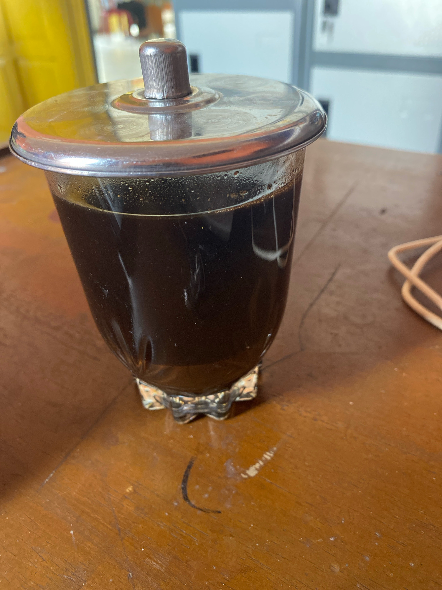 A close-up shot focuses on a clear glass filled with dark liquid, likely coffee, sitting on a wooden table. The glass has a ridged, metallic lid with a knob on top. The liquid is dark and opaque, with some fine particles visible at the surface. The wooden table surface shows signs of wear, with visible scratches and dust. In the background, a blurry yellow object and a white wall with a window can be seen. The image does not provide enough visual information to determine its location in Mojo Pahit, Indonesia.