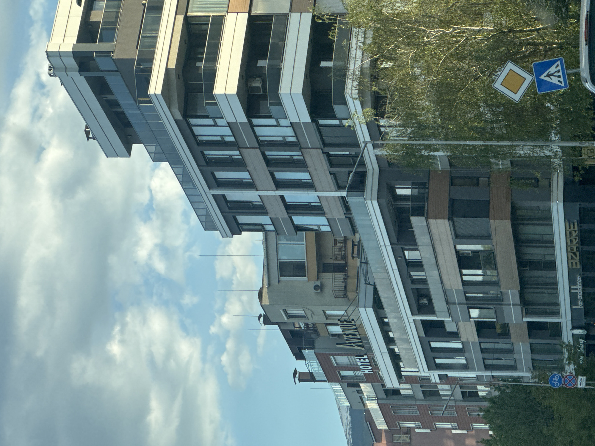 The image shows a street scene in Sofia, Bulgaria, featuring several buildings. The most prominent building is a modern, multi-story structure with a facade composed of glass and grey panels. Adjacent to it is a smaller building with the sign "HOTEL AVENUE." Further down the street, there are other buildings of varying heights and architectural styles. A street sign depicting a pedestrian crossing and a rectangular sign is visible amongst the trees. The sky, visible between the buildings, is blue with scattered clouds. The image appears to have been taken from inside a vehicle.