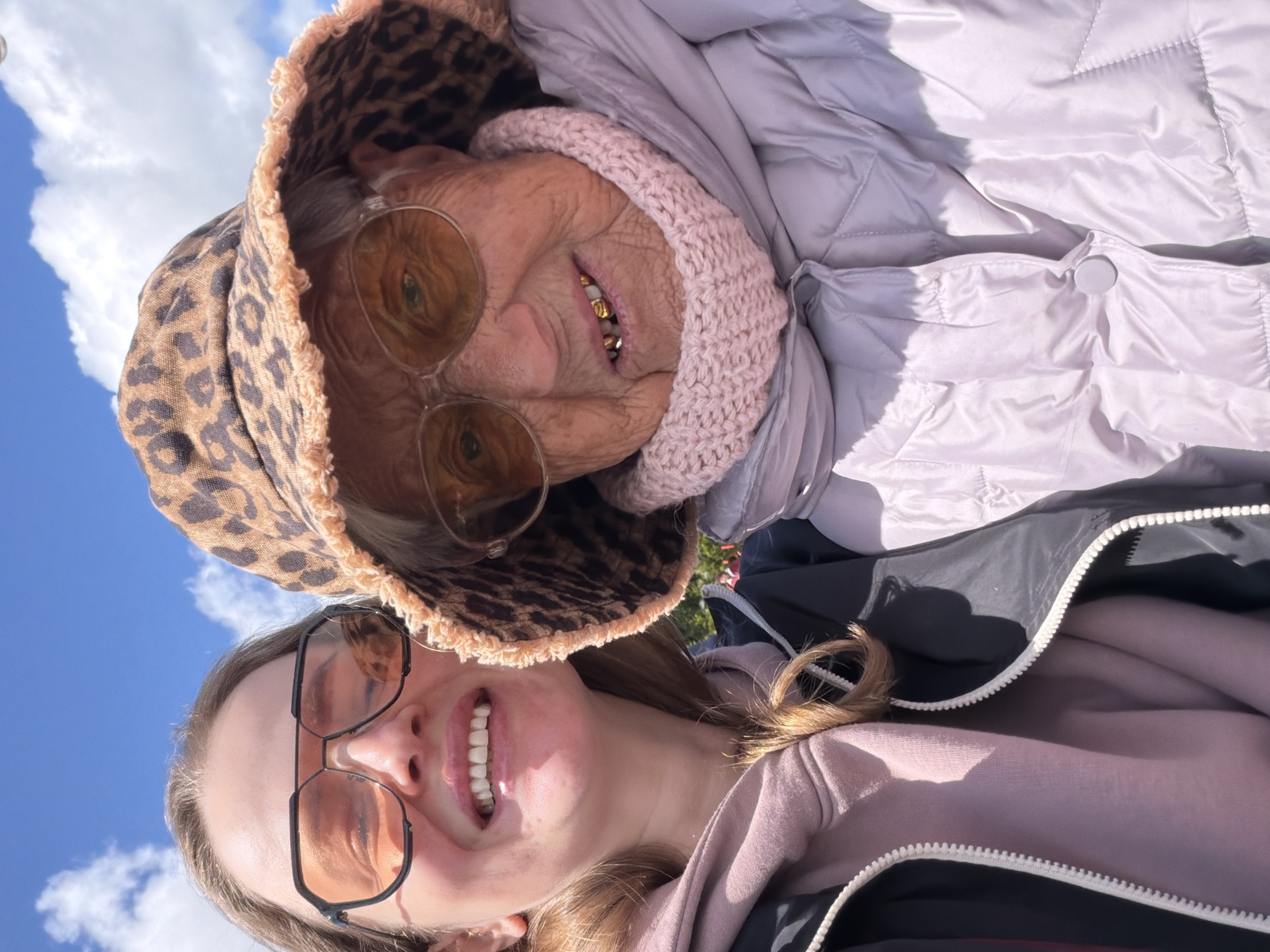 A photo taken from a low angle shows two individuals outdoors against a bright blue sky with scattered clouds, suggesting daylight. A younger woman with fair skin and shoulder-length brown hair is in the foreground wearing large square-framed sunglasses. She is smiling, revealing her teeth. She has on a pinkish hoodie with the zipper partially open and a black jacket underneath. Behind her, an older woman with wrinkled skin and a few visible gold teeth is wearing glasses with a pink, knitted cap and a leopard-print bucket hat. She also wears a light-colored, puffy coat. The setting is undefined, and the location in Valozhyn, Belarus, cannot be confirmed from visual elements.