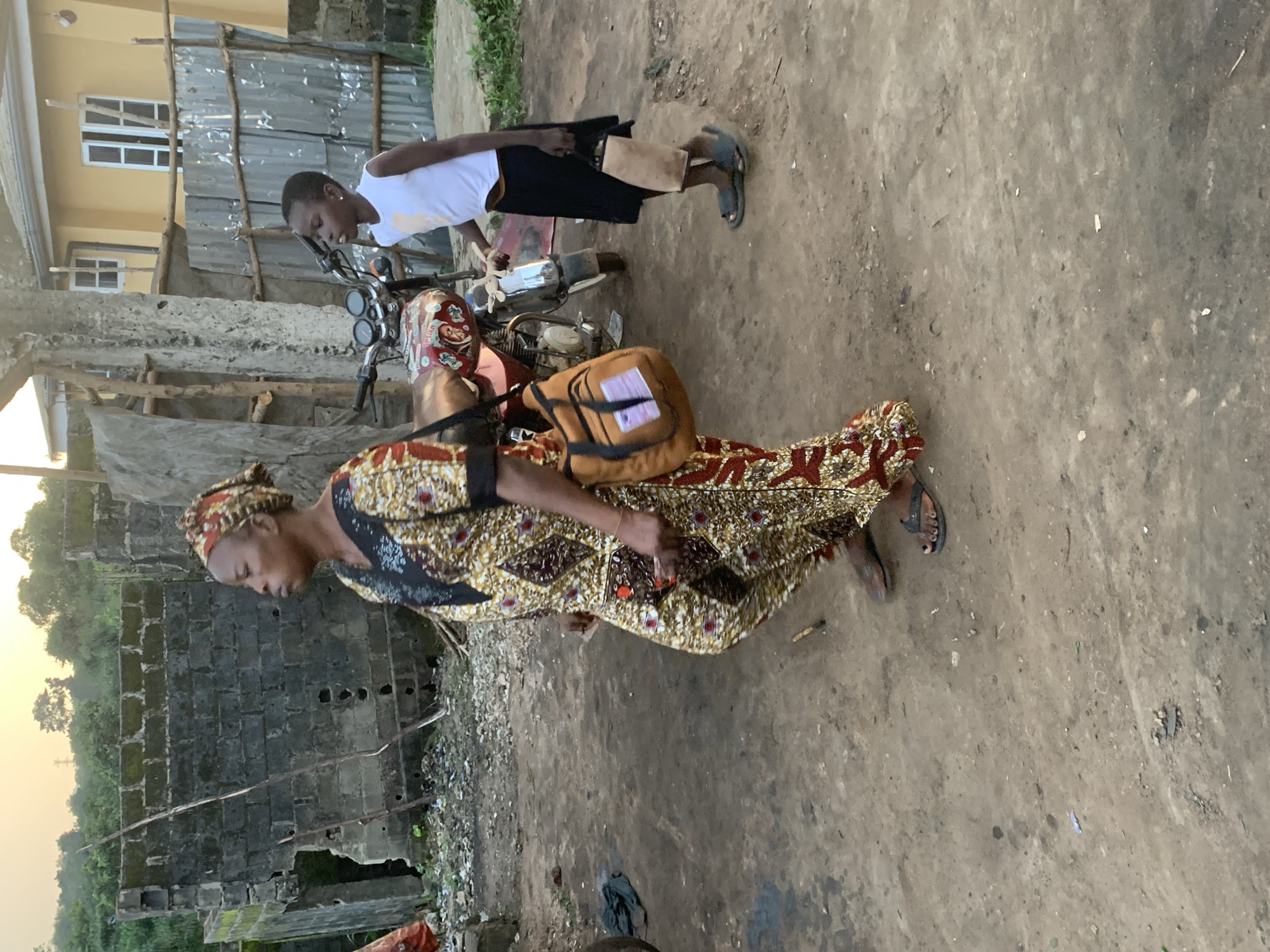 The image depicts an outdoor scene in Shagamu, Nigeria, under a hazy, pale sky. A woman, dressed in a vibrantly patterned traditional outfit and headscarf, walks towards the left of the frame. She carries a tan messenger bag slung over her shoulder. To her right, a young child, wearing a white sleeveless top and a dark skirt, stands next to a motorcycle. The child holds a light brown bag in their right hand. The motorcycle appears to be red and white, with various decals on its fuel tank. The ground is a dusty, uneven dirt surface. In the background, a partially constructed building with cinder block walls is visible, along with a yellow house with white-framed windows and a corrugated metal fence. Vegetation can be seen to the left of the cinder block structure. Debris and discarded items are scattered on the ground.
