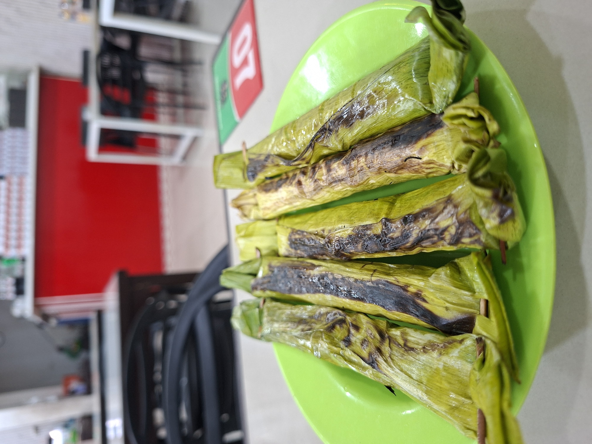 A bright green circular plate prominently displays six elongated food items in the foreground, situated on a light-colored table within an indoor dining establishment in Sigli, Indonesia. Each food item is wrapped in green leaves, likely banana leaves, exhibiting distinct dark, charred marks from grilling or heating. Small wooden skewers are visible, securing the leaf wrappers on several of the items.
The background is softly blurred, revealing a restaurant or cafe environment. Several white-topped tables paired with black chairs are discernable throughout the space. A striking red wall or counter section runs horizontally across the mid-ground. Above this red section, blurred details suggest shelves or counter space with various stacked objects, possibly bottles or containers. To the right of the plate, a portion of a green and red floor mat with the white number "20" (and potentially other characters like "BF" or "LD") is visible on the floor. No individuals are present in the frame.
