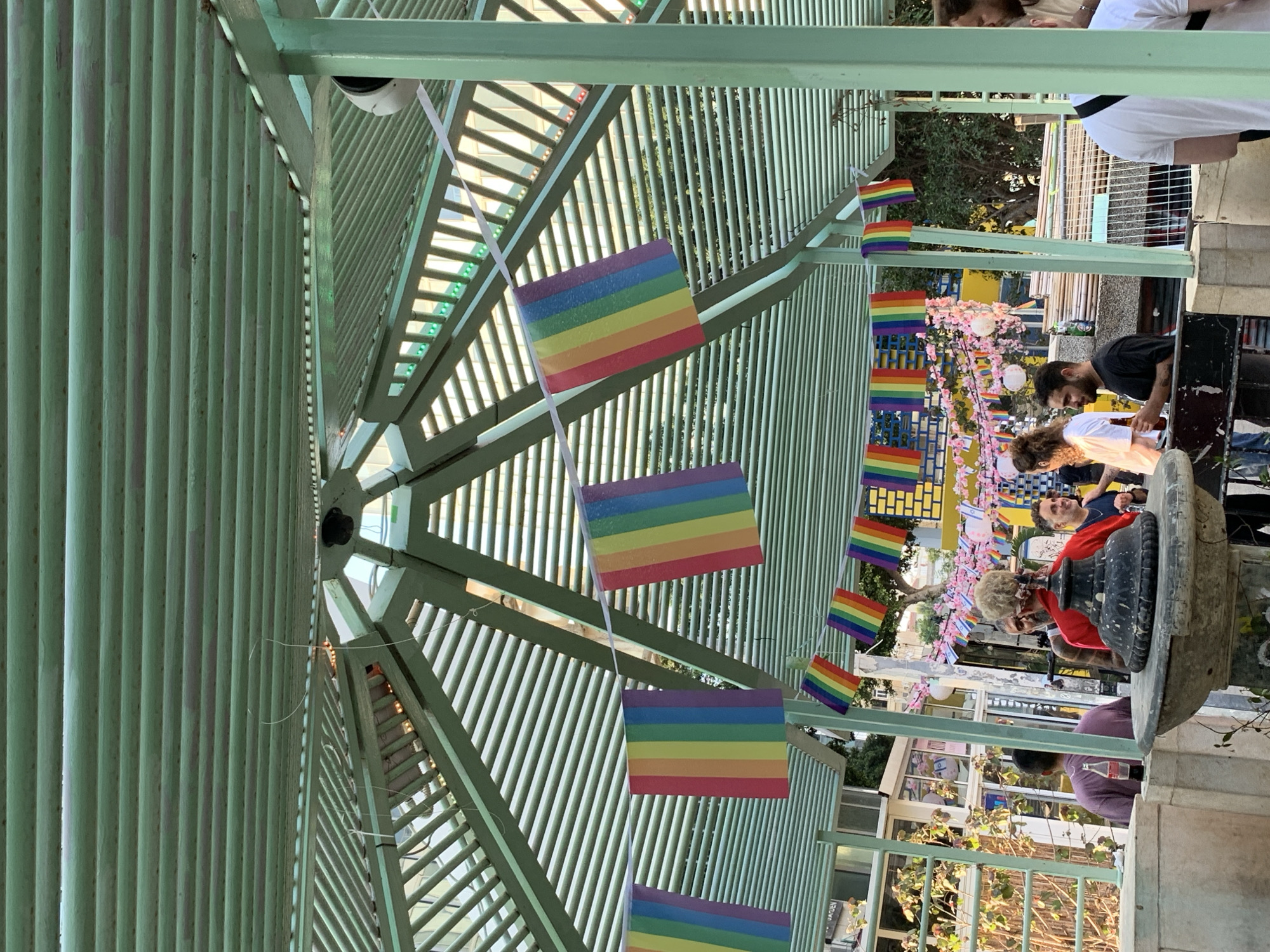 The image shows a gathering of people under a green, slatted, outdoor structure, likely a pergola or covered patio. Rainbow flag banners are strung across the structure, indicating a Pride event or celebration. In the foreground, a few individuals are visible, some engaged in conversation, others holding drinks. Behind them, there is a more densely populated area with more people and colorful decorations, including pink floral garlands and paper lanterns. The background reveals buildings and trees, suggesting an urban setting. The presence of multiple rainbow flags, Israeli flags, and a lively atmosphere point to this scene being located in Tel Aviv, Israel, during a Pride event.