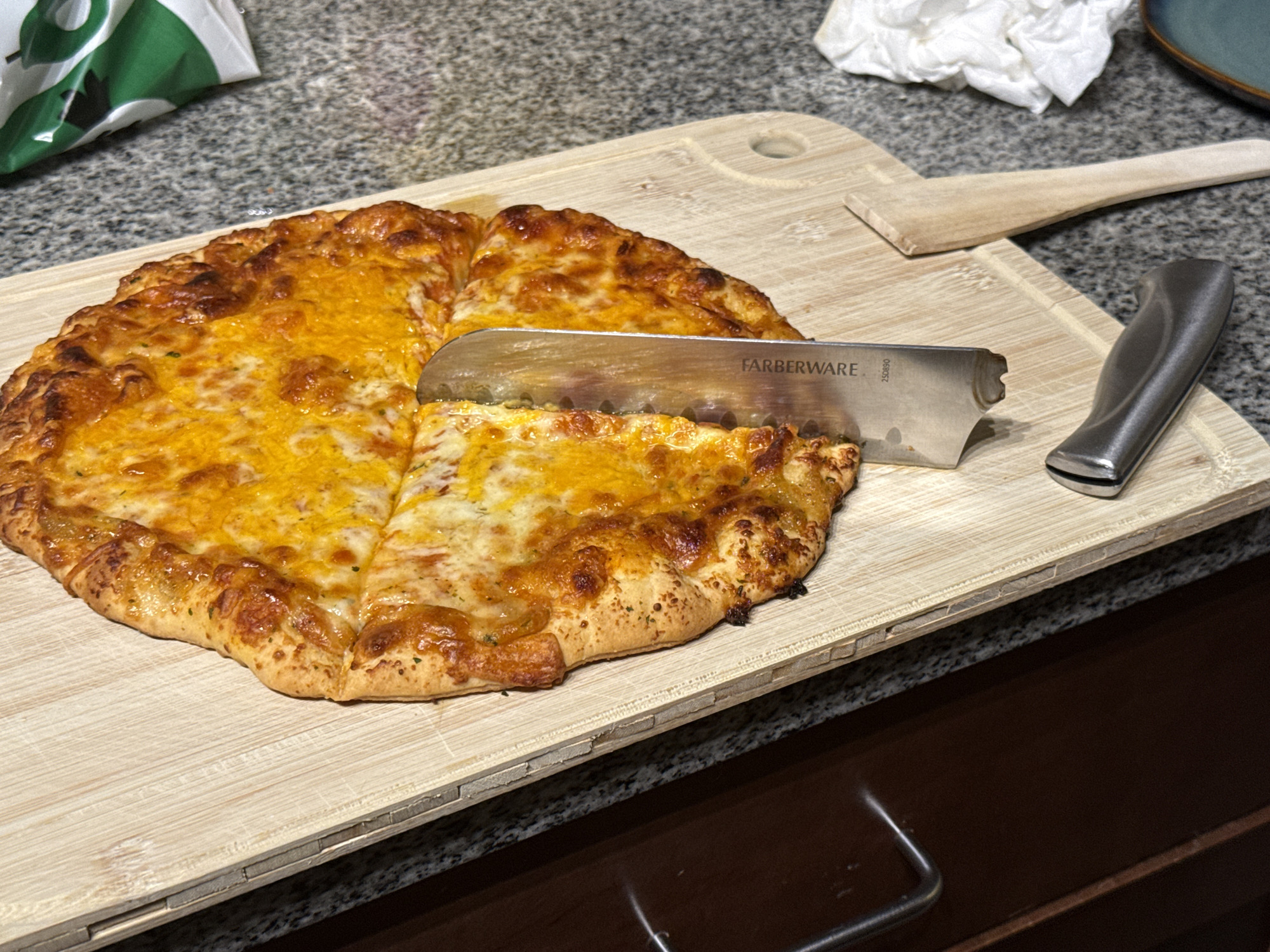 A close-up, eye-level shot shows a cheese pizza on a wooden cutting board, with a knife slicing through it. The pizza has a golden-brown crust and is covered in melted cheese, with some visible green herbs. The cutting board is light-colored wood with a light grey marbled countertop visible behind it. To the left, a crumpled white paper towel is visible, and to the far left, a portion of a green and white bag is visible. To the right of the pizza, a wooden spatula is placed on the cutting board, and a metal knife with a black handle is to the right of the pizza, appearing to be the one slicing it. The knife has the brand name "FARBERWARE" printed on its blade. The scene is indoors, with a dark wood cabinet visible in the lower portion of the image. The lighting appears to be artificial, suggesting it could be any time of day. There are no people visible in the image. The overall scene suggests a casual meal preparation or consumption.