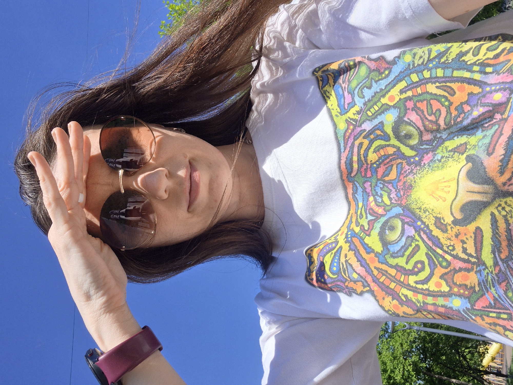 A young woman with long brown hair is pictured outdoors against a clear blue sky. She is wearing round, tinted sunglasses and a white t-shirt with a brightly colored, psychedelic tiger design. Her left hand is raised near her face, shielding her eyes from the sun. A burgundy smartwatch is visible on her left wrist. Green trees are partially visible in the background. The location and city are not discernible from the image.