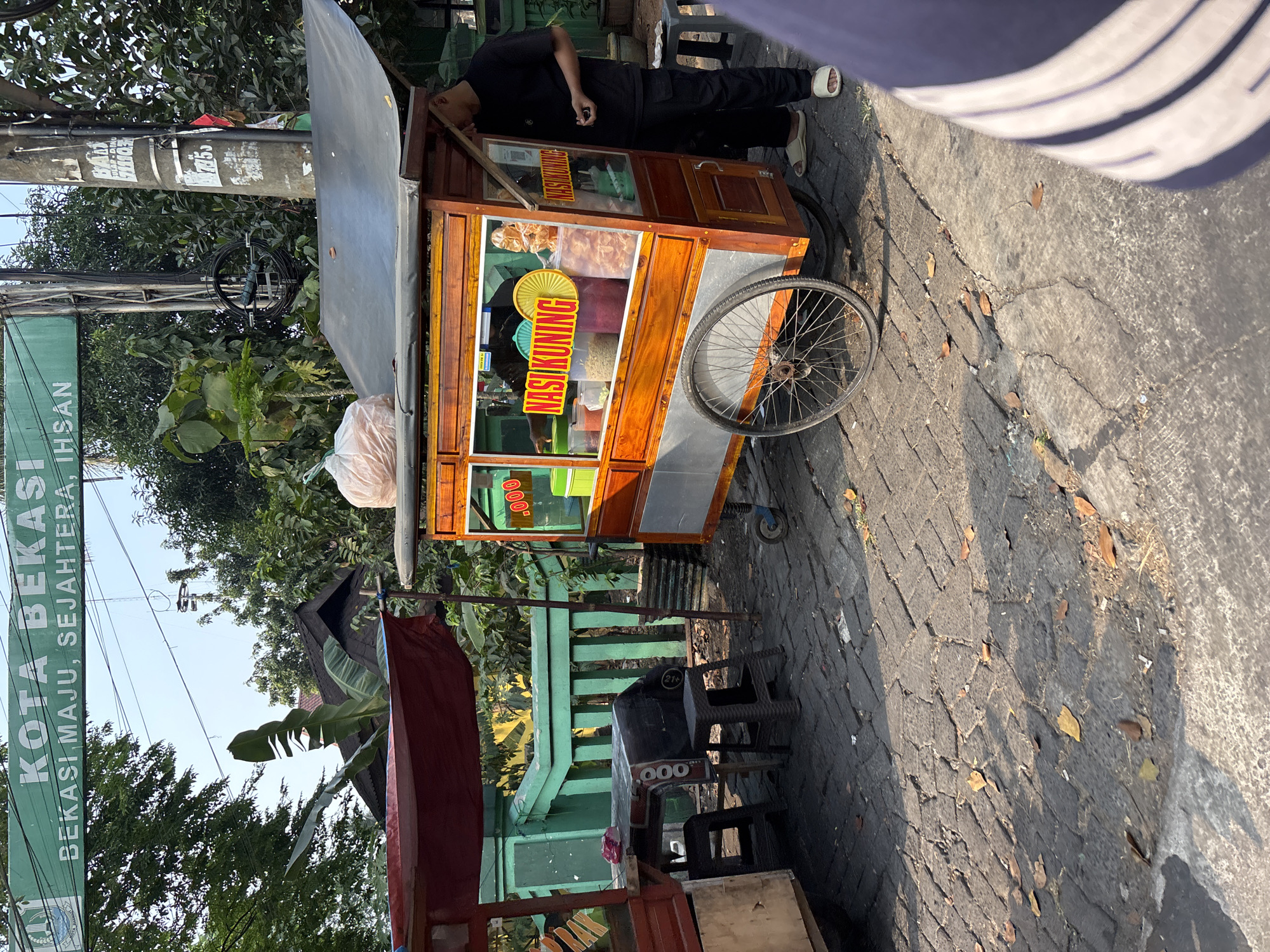 An outdoor street scene in Bekasi, Indonesia features a food cart selling "Nasi Kuning" (yellow rice). A person stands to the right of the cart, wearing dark clothing and light-colored sandals, holding a small object in their right hand. The cart itself is wooden and orange with glass windows displaying various food ingredients. To the left, another food cart with a red awning and a table with chairs is partially visible. In the background, a green fence and lush trees are present. Overhead, a green sign reads "KOTA BEKASI BEKASI MAJU, SEJAHTERA, IHSAN." The ground is paved with dark bricks and scattered with fallen leaves.