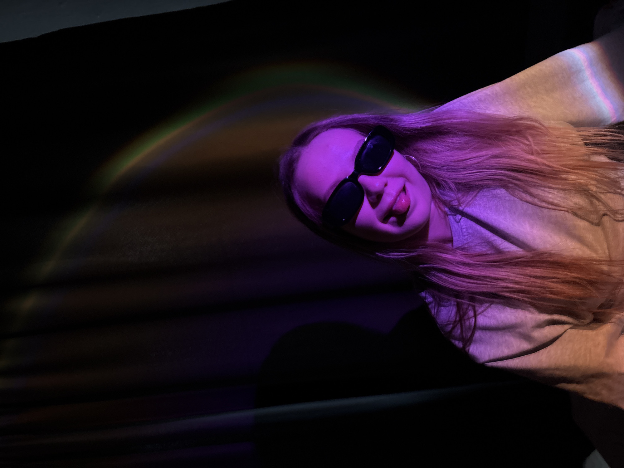 This media file captures a close-up of a young person, likely female, with long, light-colored hair, against a dark, indistinct background. The subject is wearing dark, rectangular sunglasses and a light-colored long-sleeved top. Her head is tilted, and she is smiling playfully, with her tongue slightly sticking out.
The scene is dramatically lit by artificial lighting. A strong purple and pink light illuminates the subject's face and parts of her hair and clothing. A distinctive rainbow-like light effect, resembling a prism or spectrum, forms an arc across the upper left portion of the dark background. This colorful light also subtly washes over the subject's hair and shoulder, creating streaks of orange, yellow, and blue hues. The overall setting appears to be an indoor environment or a night-time event with special lighting installations, as indicated by the dark background and vibrant, non-natural light sources. There is no visible text in the image. The playful expression of the subject, combined with the dynamic lighting, suggests a fun and casual moment.