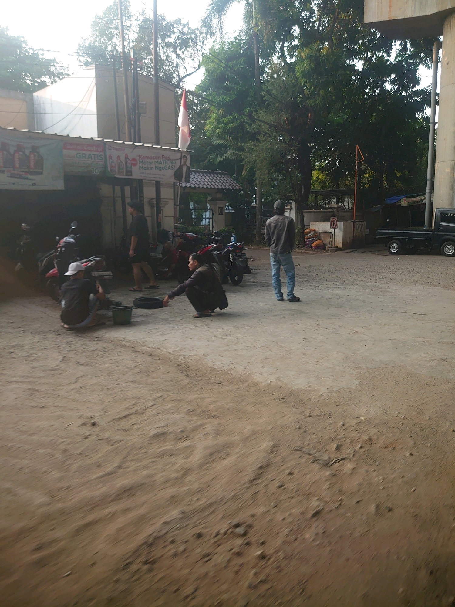 In the South Jakarta, Indonesia city location, an outdoor scene unfolds in a dusty, unpaved area, possibly a workshop or parking lot, adjacent to buildings and beneath an overpass. Three individuals are visible. In the foreground, two men are crouched down. One, wearing a white cap, is facing a green bucket and appears to be working on a tire. The other man, wearing dark clothing, is reaching towards a detached tire. Further back, a third man in a grey jacket and blue jeans stands facing away from the camera, looking towards the right side of the frame. Various motorcycles are parked to the left, suggesting a mechanic or repair service. A banner with text and images of oil bottles hangs from the building. The environment includes trees, electrical poles, and a dark-colored pickup truck parked in the background to the right. The lighting suggests it is daytime, with sunlight casting shadows and highlighting the dusty ground.