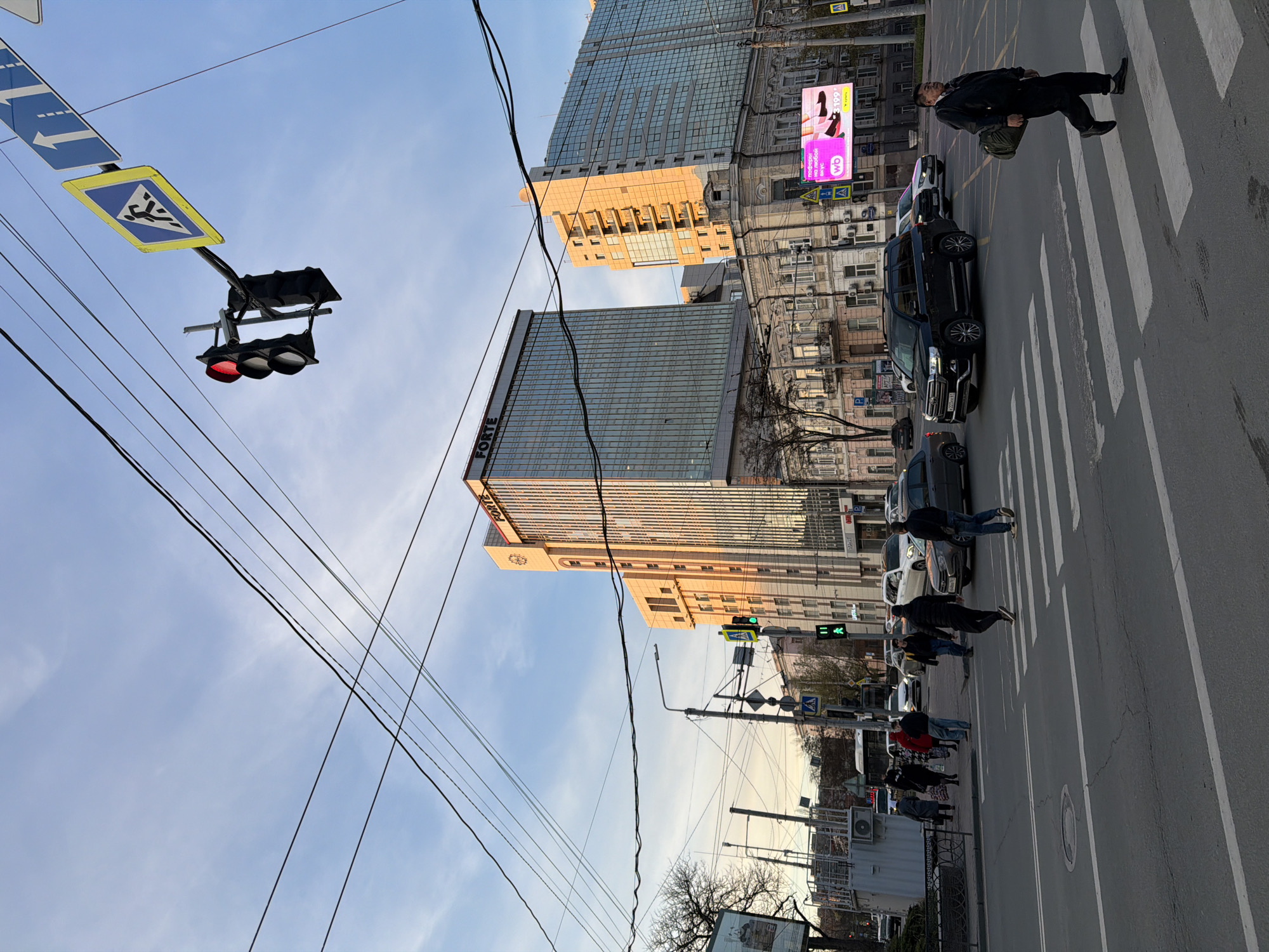 The image captures a street scene in Rostov-on-Don, Russia. Several people are using a pedestrian crossing. A black SUV is stationary on the zebra crossing. Traffic lights indicate both red and green signals on different sides of the road. In the background, there are tall buildings, including one with the "FORTE" sign visible. Power lines crisscross the sky, which is light blue. There's also an advertising billboard on the building. The pedestrian crossing is marked by standard yellow and blue pedestrian crossing signs.