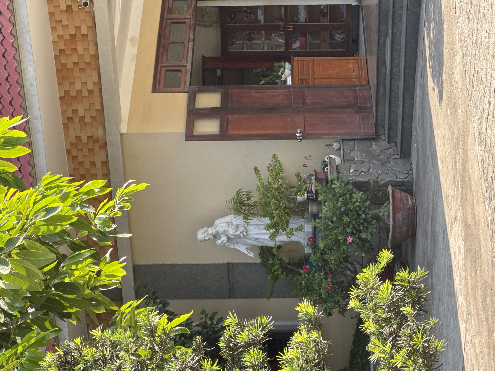 The image depicts the entrance of a building in Nha Trang Ward, Vietnam, with a focus on a white statue of a man holding a child, positioned in front of the building. The statue is surrounded by potted plants and flowers, some with pink blossoms. To the right of the statue, a building features two large wooden doors, one of which is open, revealing shelves and cabinets inside. The building's exterior is painted a light yellow, with a textured brick-like pattern above the entrance. Green foliage from trees hangs over the scene from the top left. The ground in front of the building is paved with concrete. The overall scene is illuminated by sunlight, casting shadows on the ground.