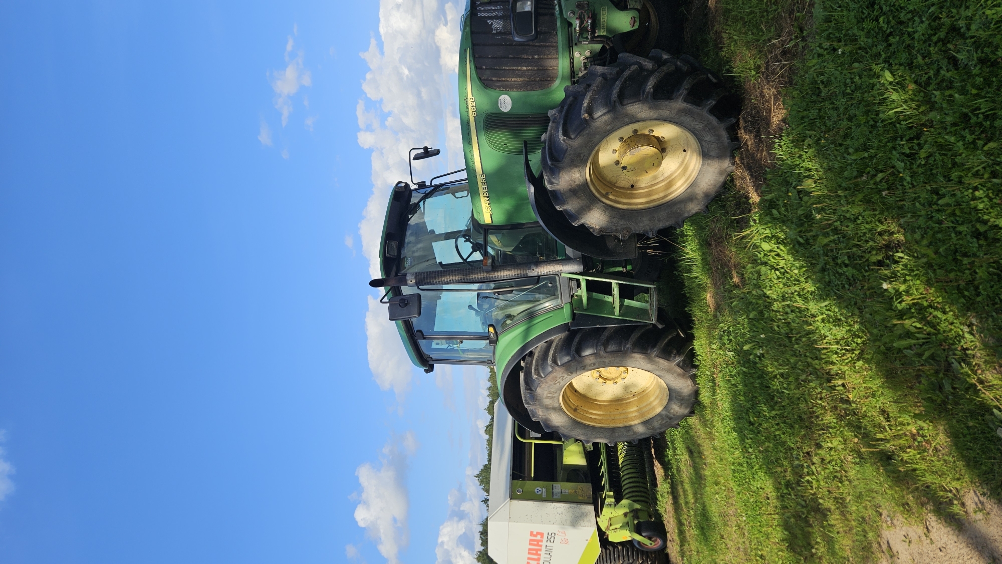 A John Deere 6620 tractor, predominantly green with yellow accents on its wheels, is parked on a grassy field under a bright blue sky with scattered white clouds. The tractor is connected to a white Claas baler, which has text reading "CLAAS ROLLANT 255" and "RS45" visible. The tractor's tires are large and black, and the sun casts shadows on the grass. In the background, a line of trees is visible, suggesting a rural or agricultural setting. The image appears to have been taken outdoors during the daytime. There is no indication of the location being in Kapūņi, Latvia.