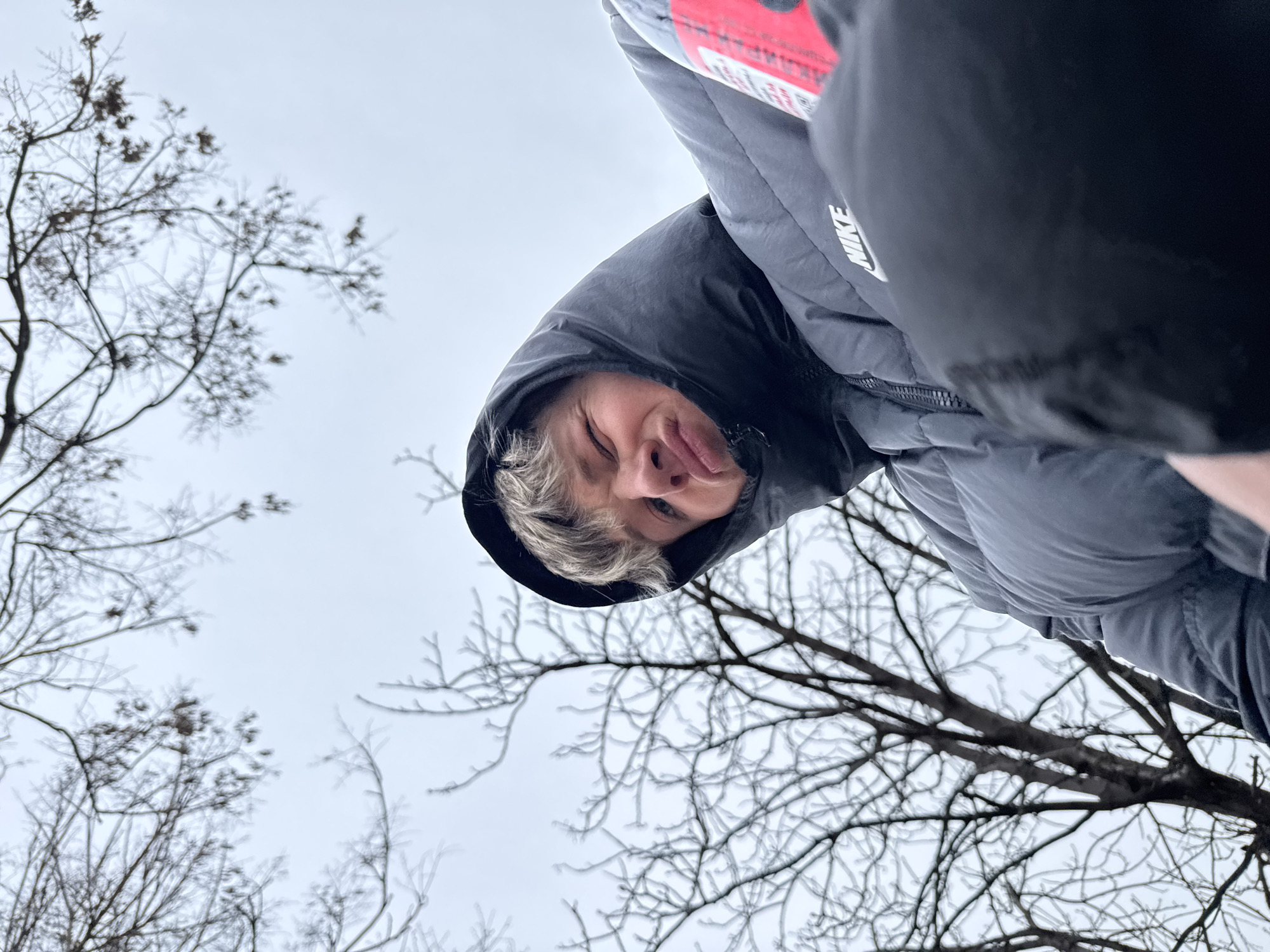 A low-angle, upside-down shot shows a person with blonde hair wearing a dark hooded Nike jacket. The person's face is visible, and they are sticking out their tongue. The background consists of bare tree branches against a gray sky. The location is likely in an outdoor area with trees in Plovdiv, Bulgaria.