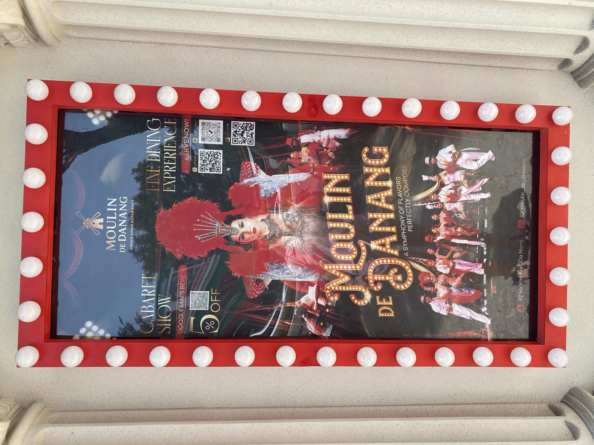 The image shows a poster for "Moulin de Danang" in Đà Nẵng, Vietnam. The poster is framed by red borders and illuminated by white bulbs. The poster features a woman in a feathered headdress and a sequined costume at the center, with a group of performers in the background. Text on the poster advertises a "Cabaret Show" and "Fine Dining Experience," with QR codes for reservations and a website. The bottom of the poster includes the address "92 Vo Van Kiet, Da Nang" and an Instagram handle "@moulin_de_danang." The poster is displayed outdoors, mounted on a building facade.