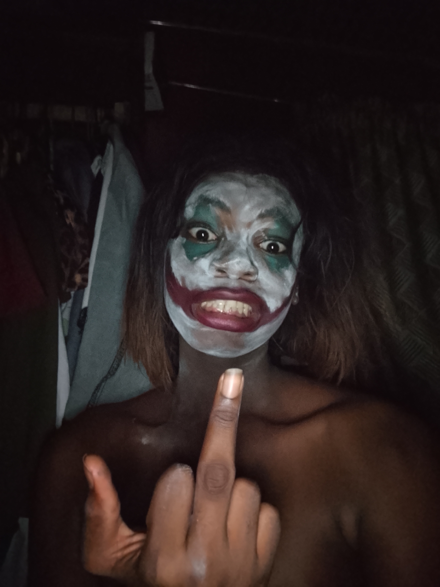 A person with dark skin is depicted from the chest up, centered in a dimly lit, indoor setting. Their face is painted in a style resembling the Joker character, featuring a white base, a wide, exaggerated red smile extending past the natural mouth line, and dark green around the eyes, which are wide open. Their dark hair frames their face.
The individual is looking directly at the camera with an intense gaze, their mouth slightly open, baring teeth in a smile. They are holding up their right hand, with the middle finger prominently extended towards the viewer in a defiant gesture. Their left hand is also visible, resting near their chest. The person appears to be topless, exposing their shoulders and upper chest.
The background is dark and indistinct, suggesting a dimly lit room or closet. Hanging clothes or fabric can be faintly discerned behind them, particularly on the left side of the frame. The overall lighting is low, with the subject's face being the most illuminated part of the image. No text is visible.