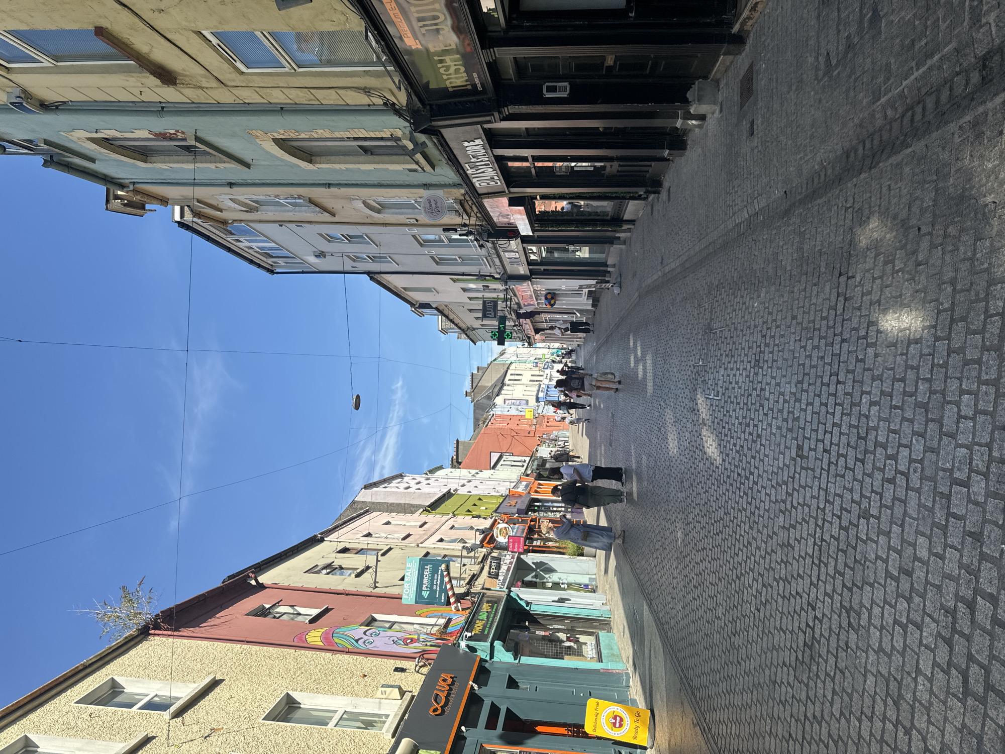 The image depicts a narrow, cobblestone street in Waterford, Ireland, lined with colorful buildings housing various shops. The sky is a clear blue with a few wispy clouds. The buildings are multi-story, with diverse architectural styles and vibrant facades, including yellow, pink, blue, green, and orange. Several shops have prominent signage. On the left side, "O'LUCA" and "PHONE AND VAPE" are visible. Further down, a "FOR SALE" sign with "PURCELL PROPERTIES" is displayed. On the right side, a sign reads "IRISH E-JUICE" and another "BLAST STORE." Several individuals are walking along the street, appearing to be shoppers or passersby. Some are in small groups, while others are walking alone. The cobblestone pavement is a prominent feature of the street. The overall scene suggests a lively, commercial street in an Irish city.