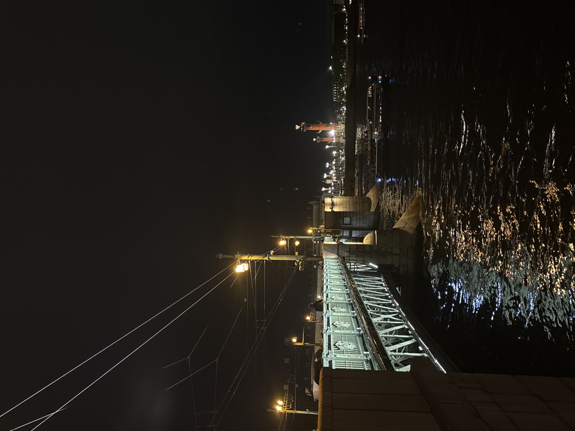 At night in Saint Petersburg, Russia, a bridge illuminated by streetlights stretches over a body of water reflecting the lights. In the distance, brightly lit Rostral Columns stand out.