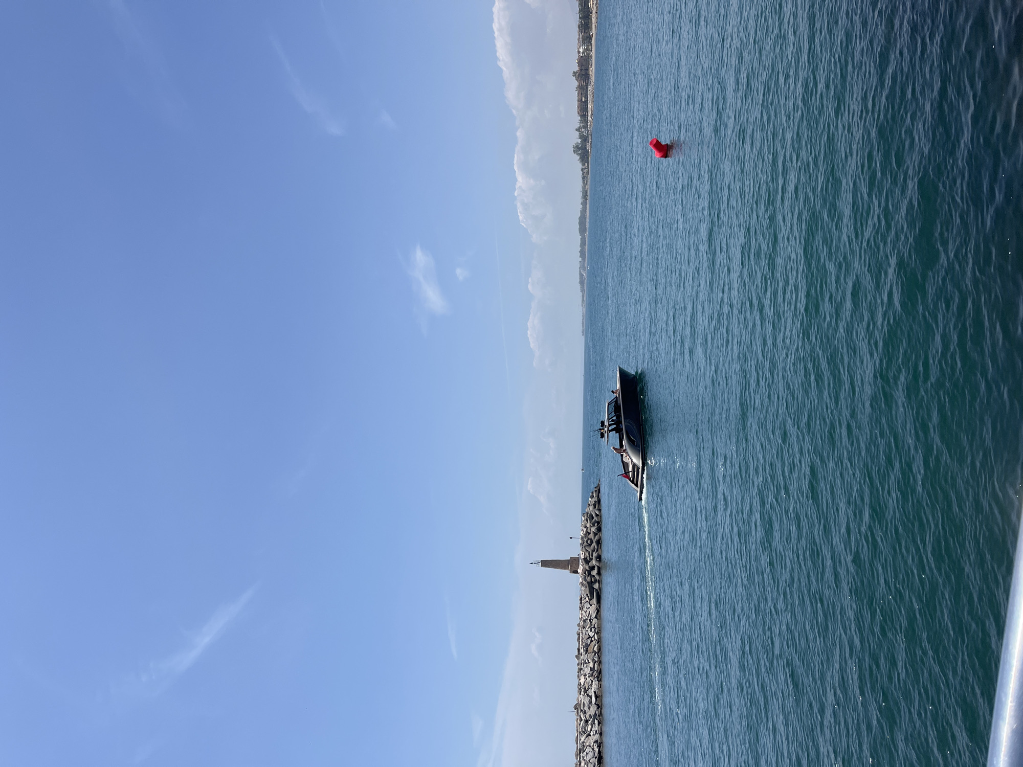 A boat is sailing in the sea toward a breakwater with a light on it. The scene appears to be off the coast of Nueva Andalucía, Spain, identifiable by the red buoy and the coastline in the background. The sky is blue with wisps of clouds.