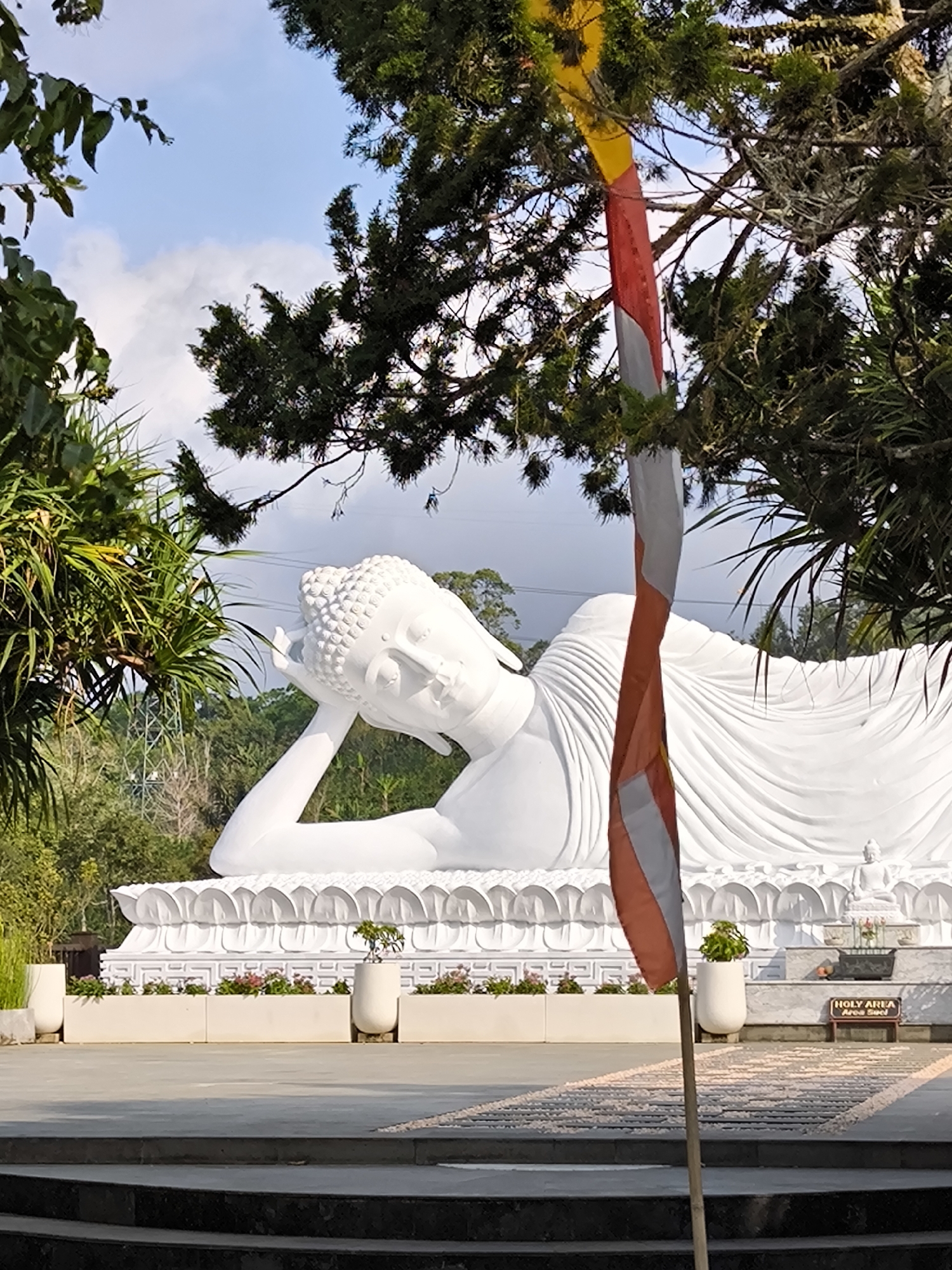The image depicts a large, white, reclining Buddha statue at the Vihara Dharma Giri Buddhist temple in Pupuan, Bali, Indonesia. The statue is the central focus, lying on its side with its head resting on its arm and hand. The statue is adorned with detailed folds, suggesting drapery. In the foreground, a flagpole with a red, white, and yellow flag is prominently positioned. The background reveals lush greenery, including trees and palm-like plants, characteristic of Bali's tropical environment. In the distance, a metal tower, possibly a transmission tower, is visible. The sky is blue with scattered white clouds. The ground in front of the statue is paved, with planters containing small bushes and flowers lining the base of the statue's platform. A sign that reads "HOLY AREA Area Suci" is also visible in the background. The overall scene conveys a sense of serenity and religious significance.