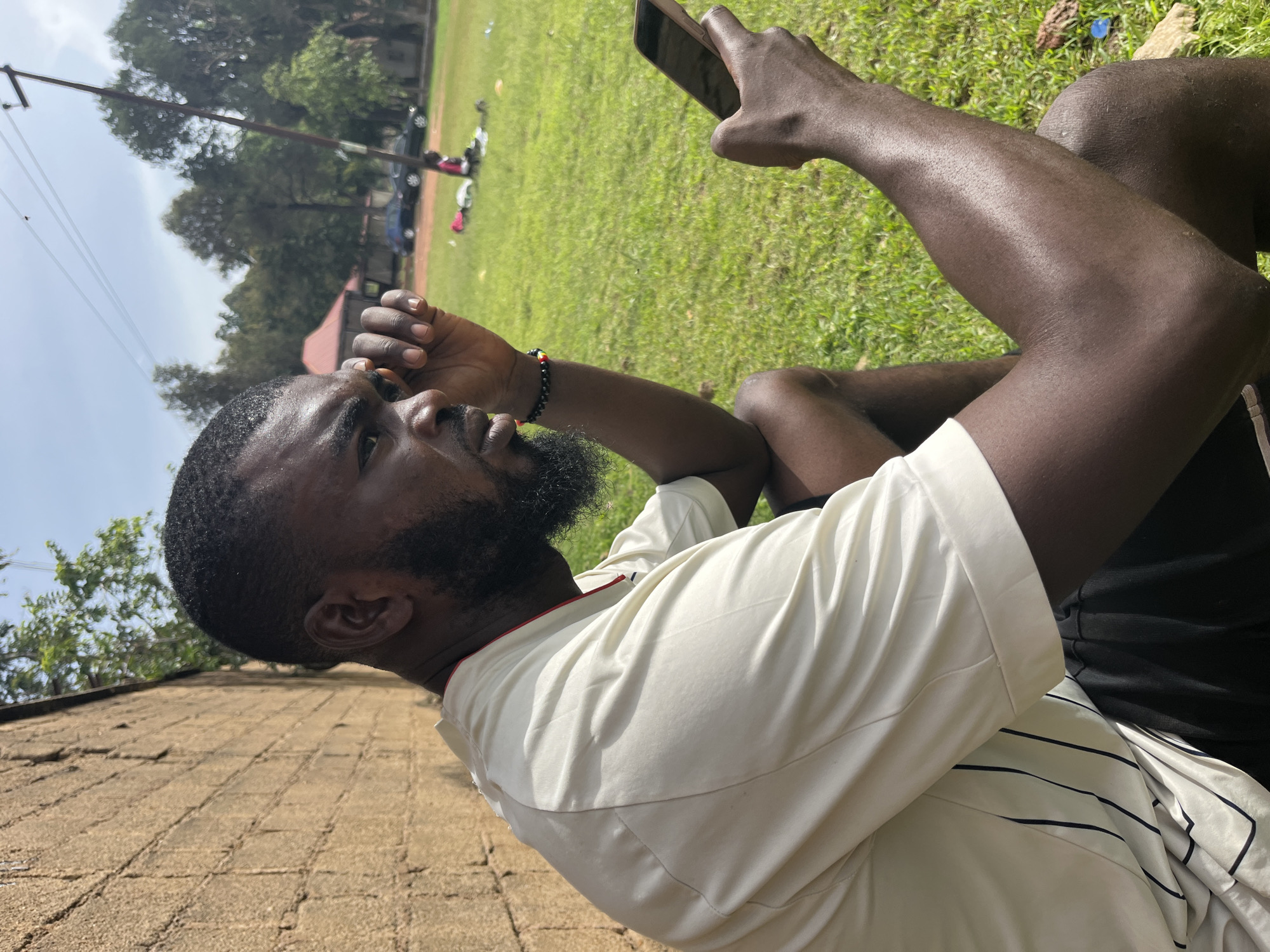 In the city of Jos, Nigeria, a Black man with a beard and dark skin sits outdoors on a grassy field. He wears a light-colored short-sleeved shirt with thin black pinstripes and dark shorts. The man is holding a smartphone in his right hand and has his left hand raised to his forehead, as if shielding his eyes or deep in thought. In the background, a low, textured brick wall runs along the left side of the frame. Beyond the wall and the grassy field, trees and buildings are visible, with a power pole and wires stretching across the sky. A person is seated in the distance on the grass, and a few cars are parked further back. The scene appears to be an outdoor recreational area or a sports field under a bright, partially cloudy sky.