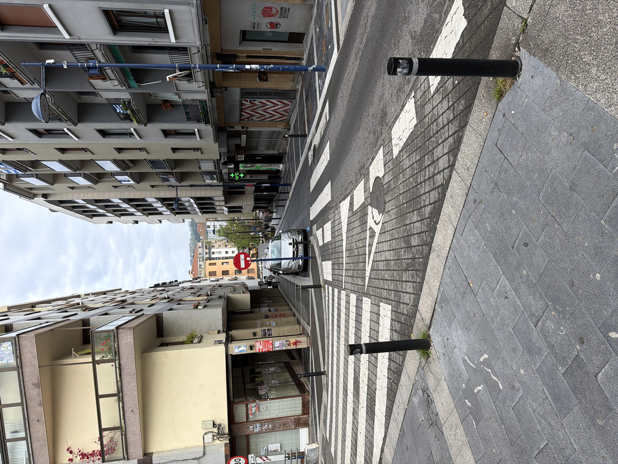 The image shows a street scene in Errenteria, Spain. The street is lined with buildings of varying heights and architectural styles. A crosswalk with white stripes and an arrow indicating the direction of pedestrian traffic is visible. Black bollards are positioned on either side of the crosswalk. A silver car is parked further down the street. A "Do Not Enter" sign is visible near the car. Street signs and indicators of commercial establishments, including a pharmacy, are visible along the street. The sky is overcast.