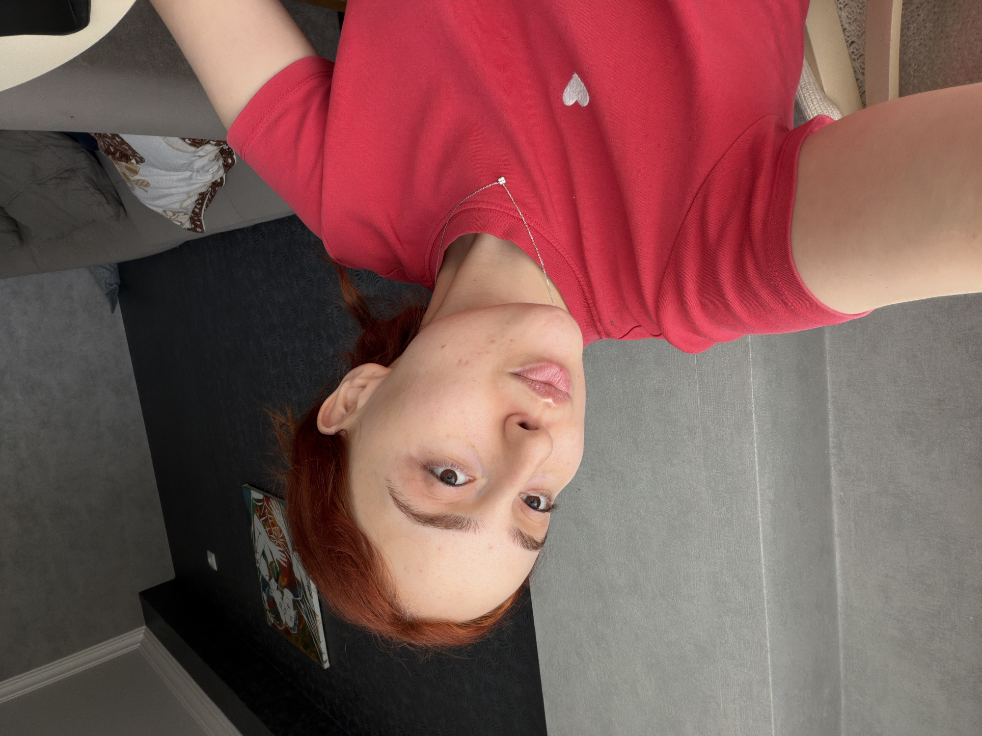 The image, originally rotated, depicts a young woman lying down and looking directly at the camera in an indoor setting. She has reddish-brown hair, styled loosely, and light skin. Her expression is neutral, with her eyes open and lips slightly parted.
She is wearing a plain red t-shirt that features a small white heart embroidered on the left chest area. A thin silver necklace with a tiny, clear, possibly floral-shaped pendant rests on her collarbone. Her arms are visible, showing bare skin.
The background behind her head, to the left (when correctly oriented), includes a dark grey or black wall with a white baseboard. A small, colorful picture or postcard, depicting abstract or stylized figures, is placed on a black shelf or ledge against this wall. Further left, a portion of a grey upholstered sofa or bed is visible, with a white and brown patterned throw pillow resting on it. To the right of her head, the background consists of a light grey wall with a subtle textured finish and faint vertical lines.
The overall setting suggests a casual home environment, possibly a living room or bedroom, with contemporary interior design elements. There is no visible text in the image. The time of day is not explicitly indicated but appears to be well-lit indoors.