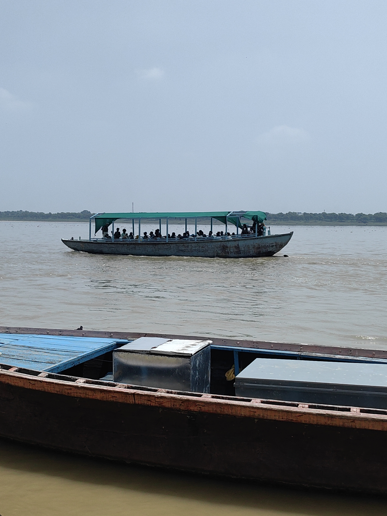 An eye-level shot captures a passenger ferry navigating the muddy Ganges River in Varanasi, India, under an overcast sky. The ferry, a long, flat-bottomed vessel with a green canopy, is filled with passengers. The boat's hull appears weathered and worn. In the foreground, the edge of another wooden boat is visible, its deck painted blue and featuring metal storage containers. The river is wide and calm, with a distant treeline bordering the horizon. The overall atmosphere is serene and suggests a typical day on the Ganges.