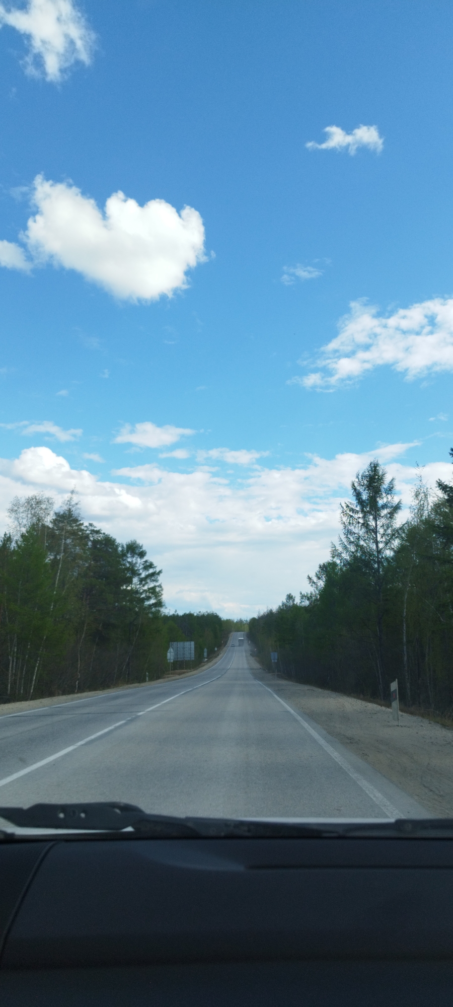 The image depicts a view from inside a vehicle, looking out onto a straight, asphalt road in Tabaga, Russia. The road stretches into the distance, flanked on both sides by green trees. A white line marks the edge of the road. The sky is predominantly blue with scattered white clouds. The interior of the vehicle, including the dashboard and windshield wiper, is visible at the bottom of the frame.