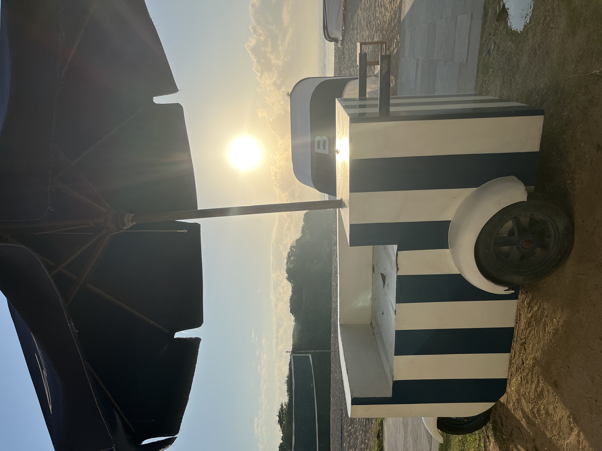 This image depicts a beach scene in Kuta Selatan, Indonesia, at sunrise or sunset. A dark blue and white striped cart, possibly a food or drink vendor, is positioned in the foreground under a large dark umbrella. The cart has two wheels visible and a counter area. Behind the cart, a volleyball net is set up on the sandy beach. The sun is low on the horizon, casting a bright glare and creating a lens flare effect. The sky is partly cloudy, and the ocean is visible in the distance. The environment suggests a resort or recreational area. There are no people visible in the image.