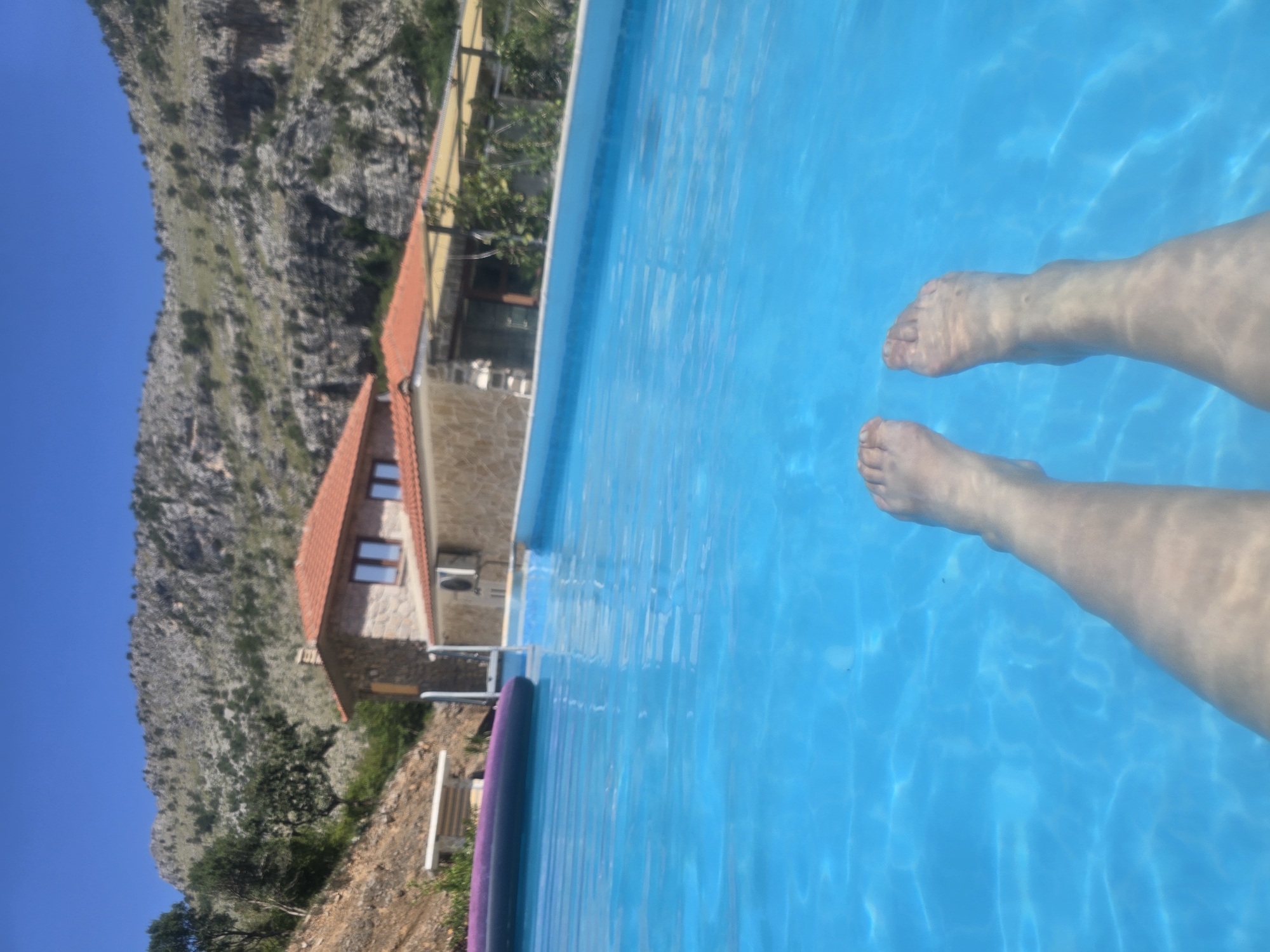 The image depicts a first-person perspective of a person's legs and feet submerged in a bright blue swimming pool. The water is clear, revealing the details of their skin. In the background, there is a stone-built house with a red-tiled roof, and beyond that, a rocky, mountainous landscape under a clear blue sky. To the left of the house, part of a purple inflatable pool float is visible. The scene suggests a sunny day in a rural or mountainous area, likely a vacation setting.
