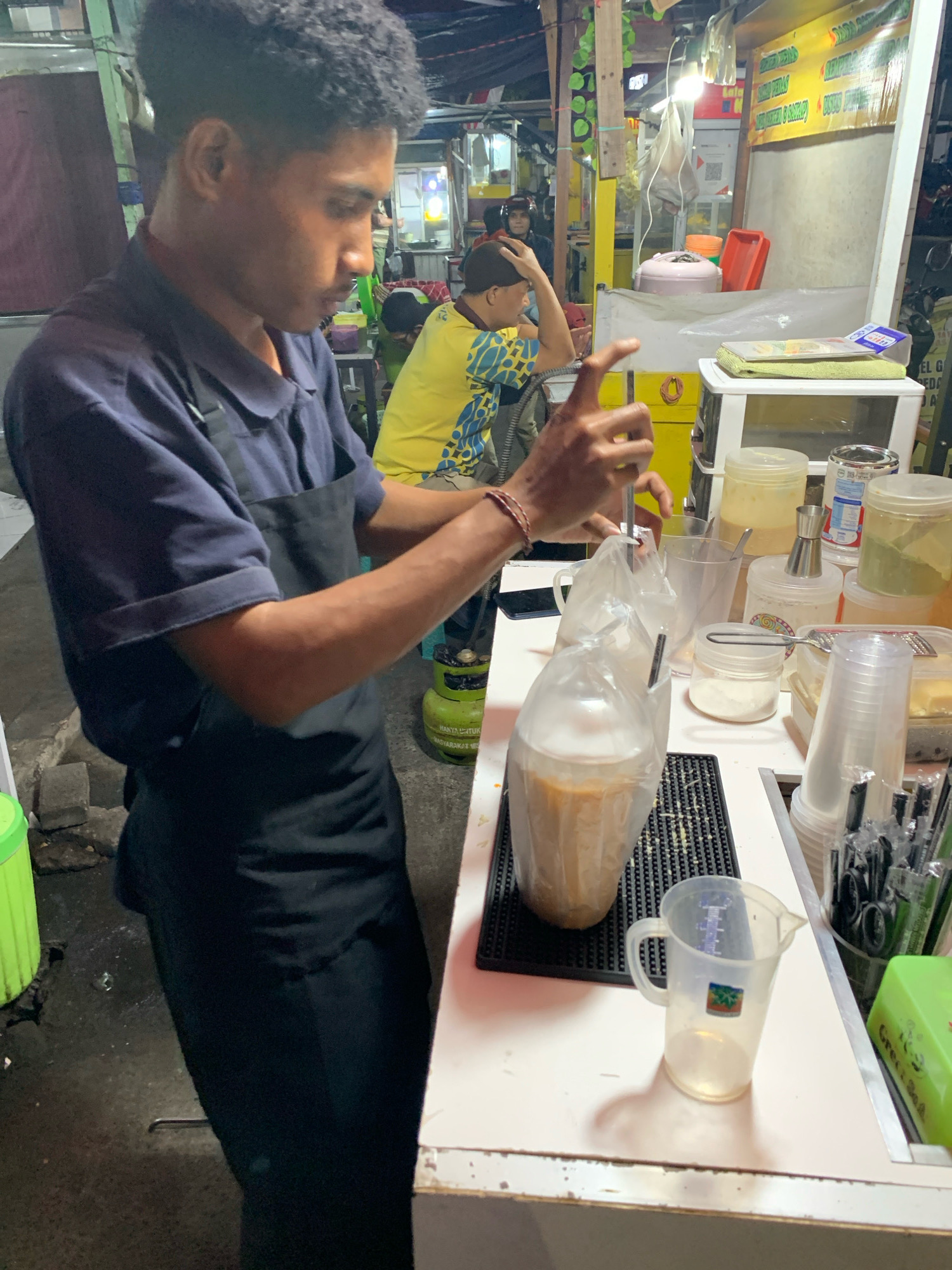 A young man wearing a navy blue polo shirt and a black apron is preparing a beverage at a street food stall. He is using a silver stirring tool inside a plastic bag containing a brown liquid with foam. The stall appears to be located in Malang, Indonesia, as suggested by the Indonesian text visible on the yellow banner above the stall and the general appearance of the surroundings, which is characteristic of street food vendors in the city.
To his left, a green gas cylinder is visible, and further in the background, other people are seated, one of whom is wearing a yellow and blue patterned shirt. The stall is equipped with various containers of ingredients, a clear pitcher, and stacks of disposable cups. The counter is cluttered with items, including a small scale, various jars, and a container of straws. The ground around the stall is wet and somewhat dirty, indicating an outdoor, bustling environment typical of urban street markets. The lighting appears to be artificial, with multiple bulbs illuminating the stall area.