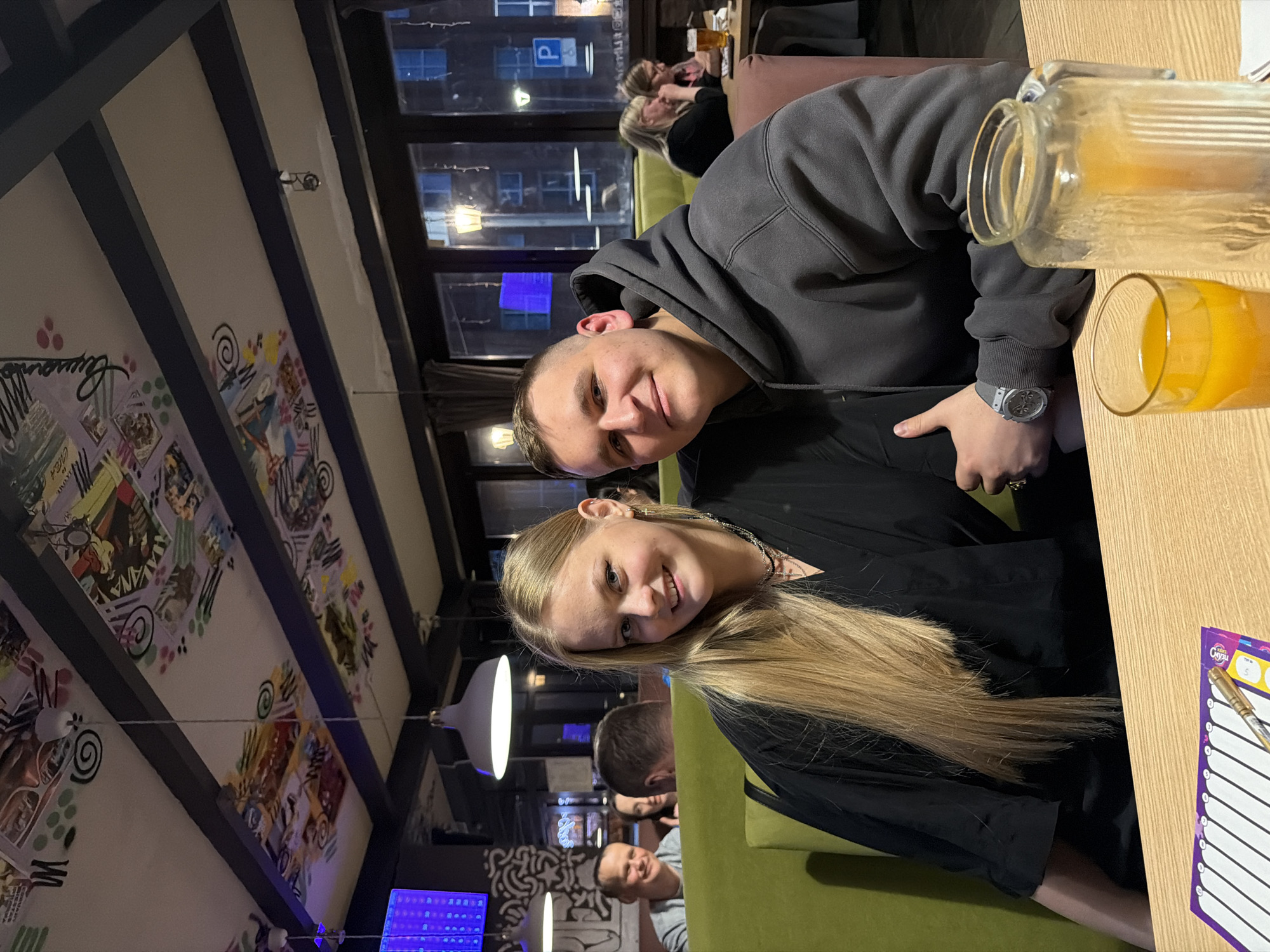 The image portrays two young individuals, a male and a female, seated at a light wooden table in an indoor setting, likely a cafe or restaurant, during the nighttime. Both are smiling and looking directly at the camera, conveying a cheerful and relaxed mood.
The male subject, on the left, wears a dark grey hooded sweatshirt and has short, light brown hair. He is seen wearing a silver-colored wristwatch on his left wrist. The female subject, seated to his right, wears a black top and has long, straight blonde hair. She has a gold-colored ring on her right hand. They are seated on green upholstered furniture.
On the table in front of them are a large glass pitcher and a glass, both filled with an orange liquid, presumably juice. A small purple-edged notepad, featuring the visible text "Супер" (Super) and horizontal lines, rests on the table next to a gold-colored pen.
The background shows large windows looking out onto an urban night scene, with visible streetlights and reflections confirming the evening hours. A blue parking sign with a stylized 'P' is discernible through the window. Further back, blurred figures of other patrons are visible, seated on brown upholstered seating. The ceiling features dark beams and panels decorated with vibrant, artistic designs, including abstract shapes, swirls, and stylized illustrations, some of which contain illegible text that appears to be in Cyrillic. Various light fixtures illuminate the space.