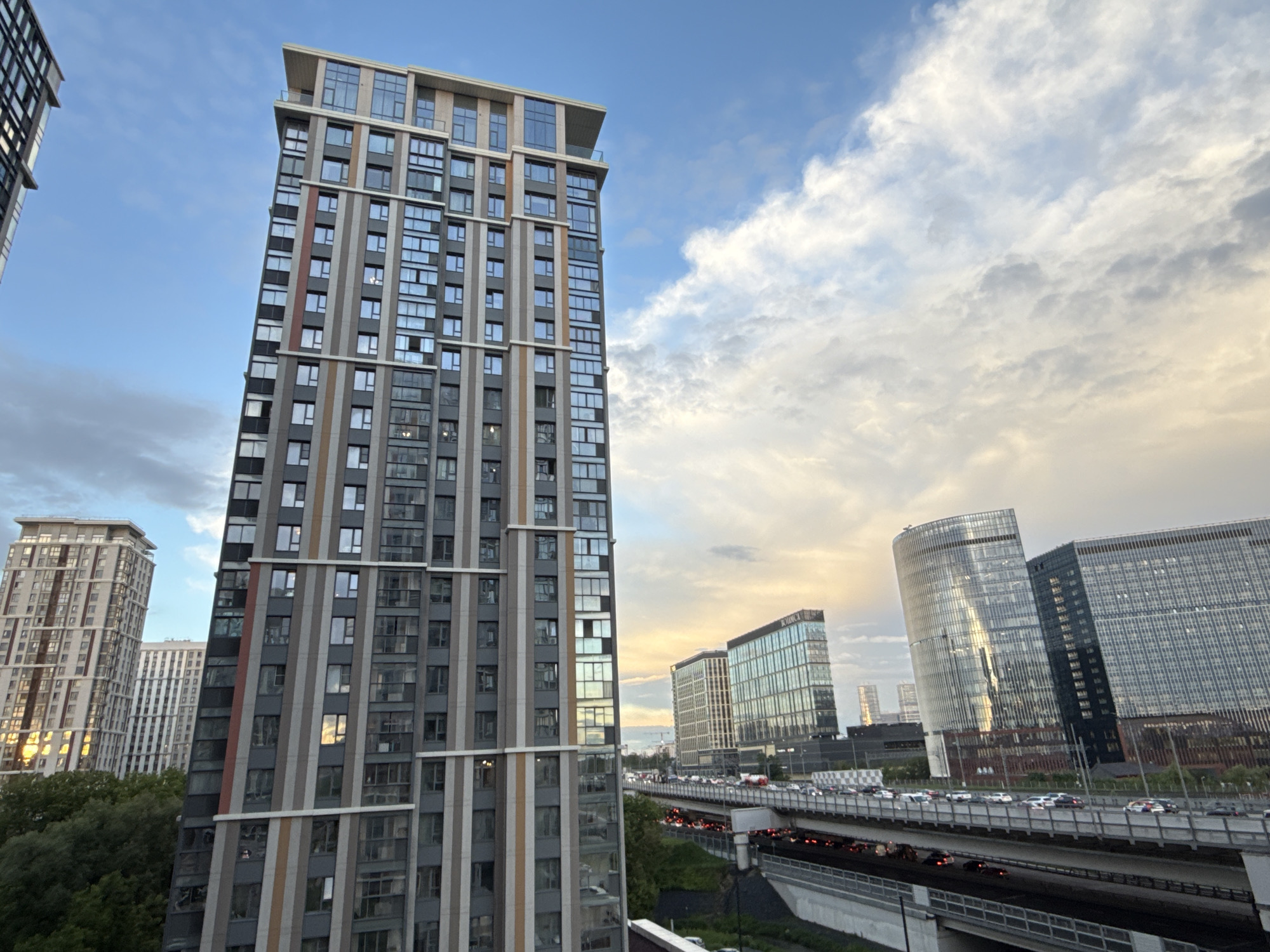 The image depicts a view of Khimki, Russia, featuring a tall, modern residential tower with a gray facade accented by vertical strips of white, brown, and red. Other similar buildings are partially visible to the left, with their facades displaying mirrored windows. A highway viaduct teeming with car traffic is in the mid-ground, and behind it, there are several office buildings, including cylindrical-shaped structures with mirrored glass facades. Above, the sky is mostly cloudy, with a gradient from light blue to a hint of orange near the horizon. The time of day appears to be late afternoon or early evening. The vantage point suggests a high-rise location, likely from another building. There are green trees visible between the building and the elevated highway.