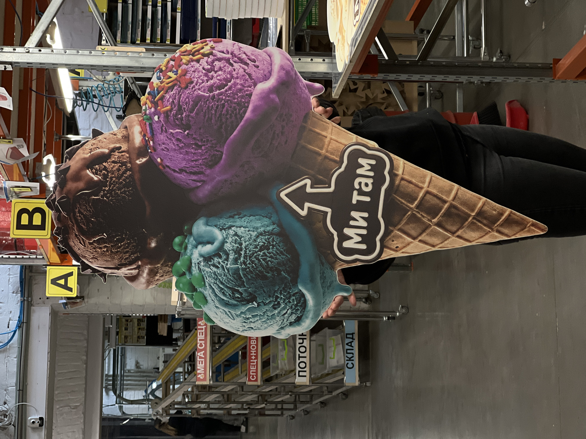 A person is seen holding a large, colorful cardboard cutout of an ice cream cone within what appears to be a warehouse or storage facility. The cutout features three scoops of ice cream: a purple scoop with colorful sprinkles on top, a chocolate-brown scoop in the middle, and a blue-green scoop with green spherical toppings at the bottom, all served in a waffle cone. An arrow pointing left with the Cyrillic text "МЫ ТАМ" (My Tam), meaning "This way" or "We are there," is prominently displayed on the cone.
The person, visible from the mid-torso down, is wearing dark clothing—a black top and black pants—and holds the ice cream cutout upright with both hands.
The background is filled with industrial metal shelving units, characteristic of a warehouse environment. Various signs and labels are attached to these shelves, also in Cyrillic. Notable labels include "МЕГА СПЕЦ" (Mega Special), "СПЕЦ+НОВЫЙ" (Special+New), "ПОТОЧНЫЙ" (Flow / Production Line), and "СКЛАД" (Warehouse). Yellow signs with black letters "A" and "B" are also visible. The area is illuminated by fluorescent lights. The floor and walls appear to be concrete or painted grey. A blue coiled hose or cable hangs from the ceiling or shelving, and a red dustpan and brush are on the floor near the person's feet.